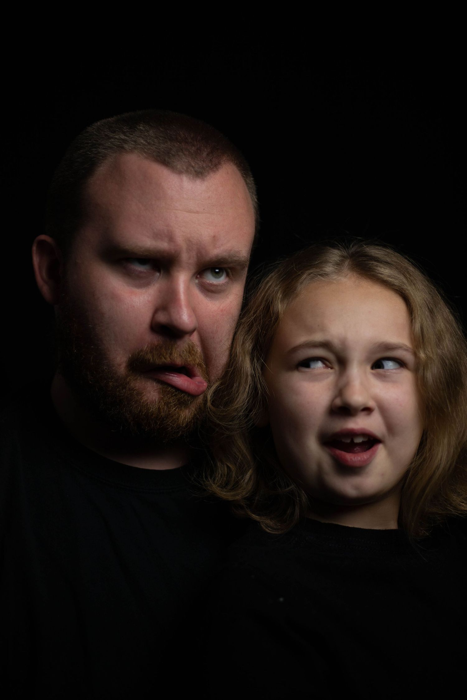 A bearded man and young blonde girl make silly faces together against a dark background.