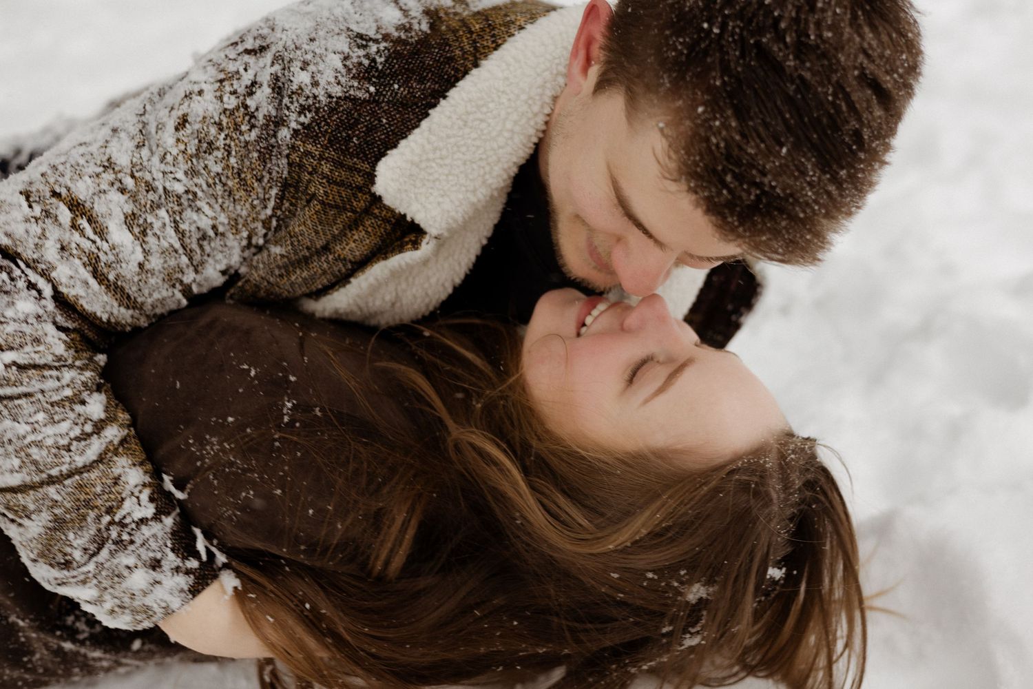A romantic winter scene with a couple lying in snow wearing cozy jackets and sharing an intimate moment.