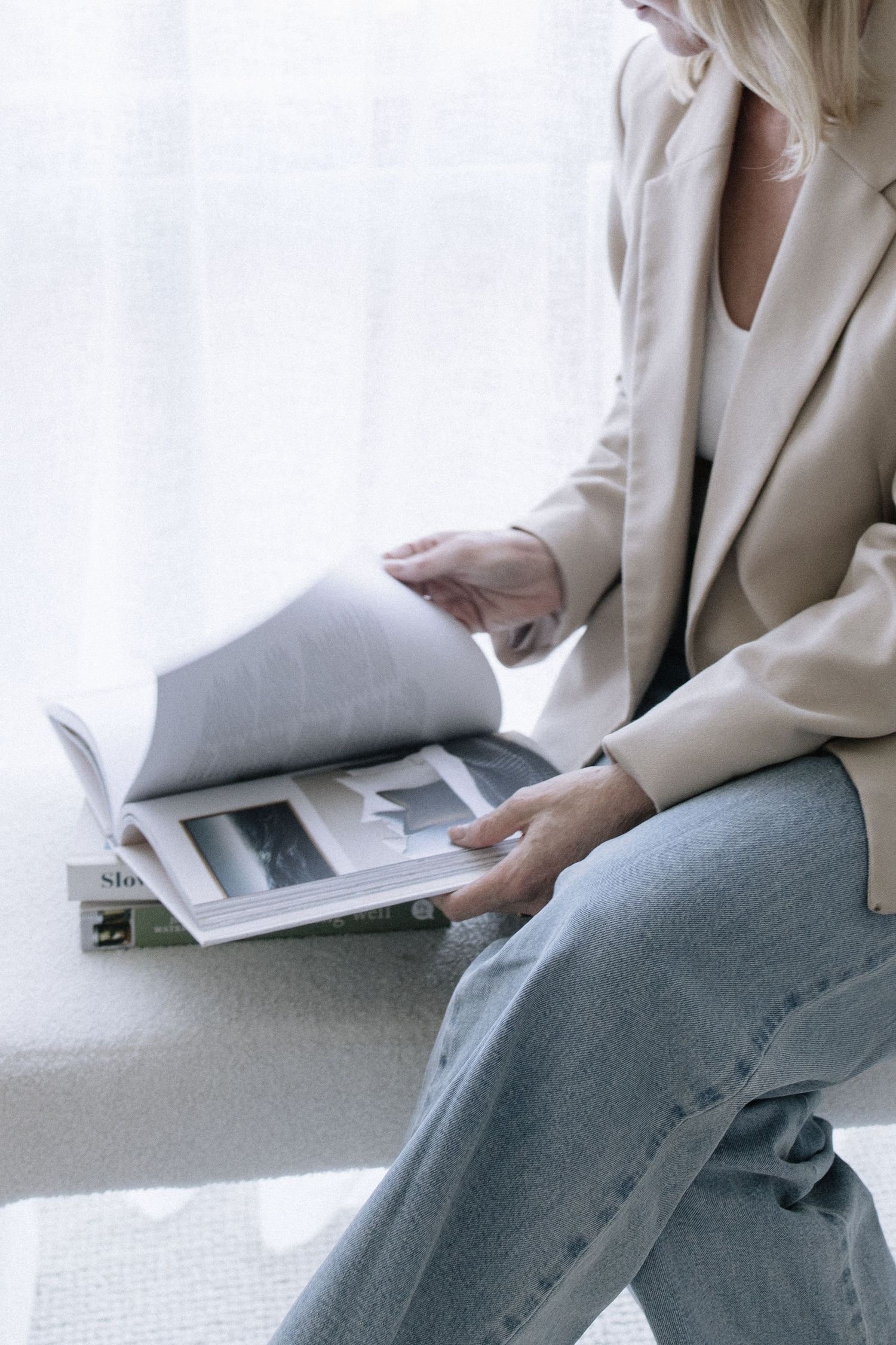 Close-up of woman in beige blazer flipping through magazine pages while seated near bright window.