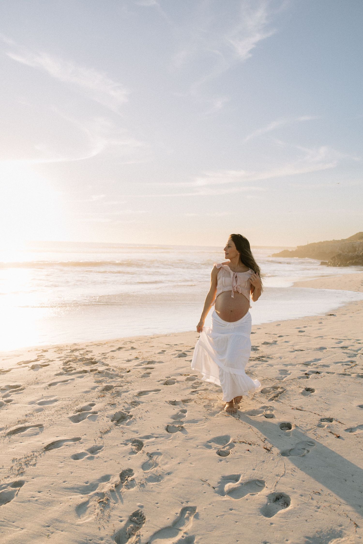 Pregnant woman in white outfit walking barefoot on sandy beach during golden sunset.