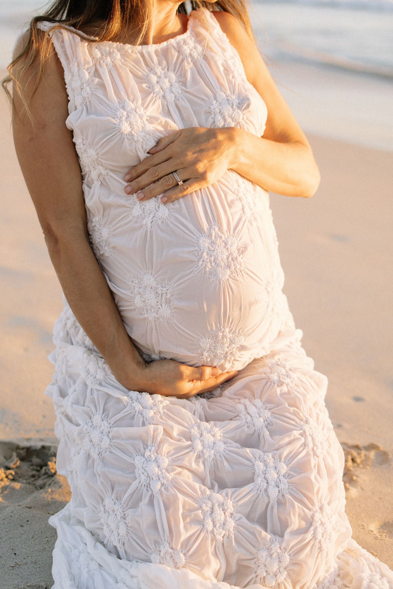 A pregnant woman in a white floral textured dress cradles her bump while kneeling on a sandy beach.