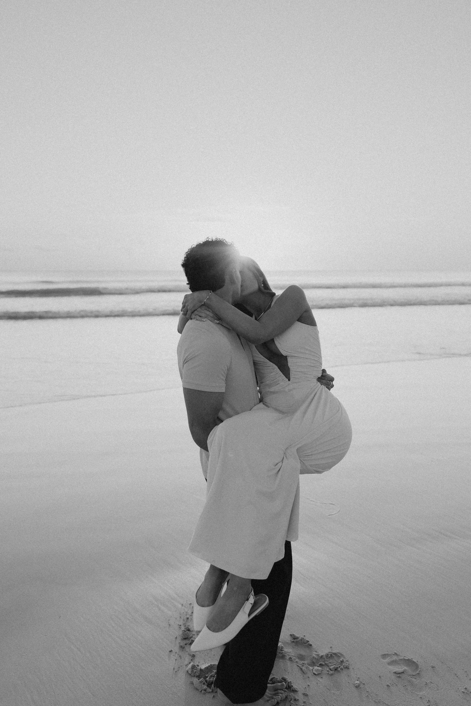 Black and white image of couple kissing and embracing on beach at sunset with glowing backlight.