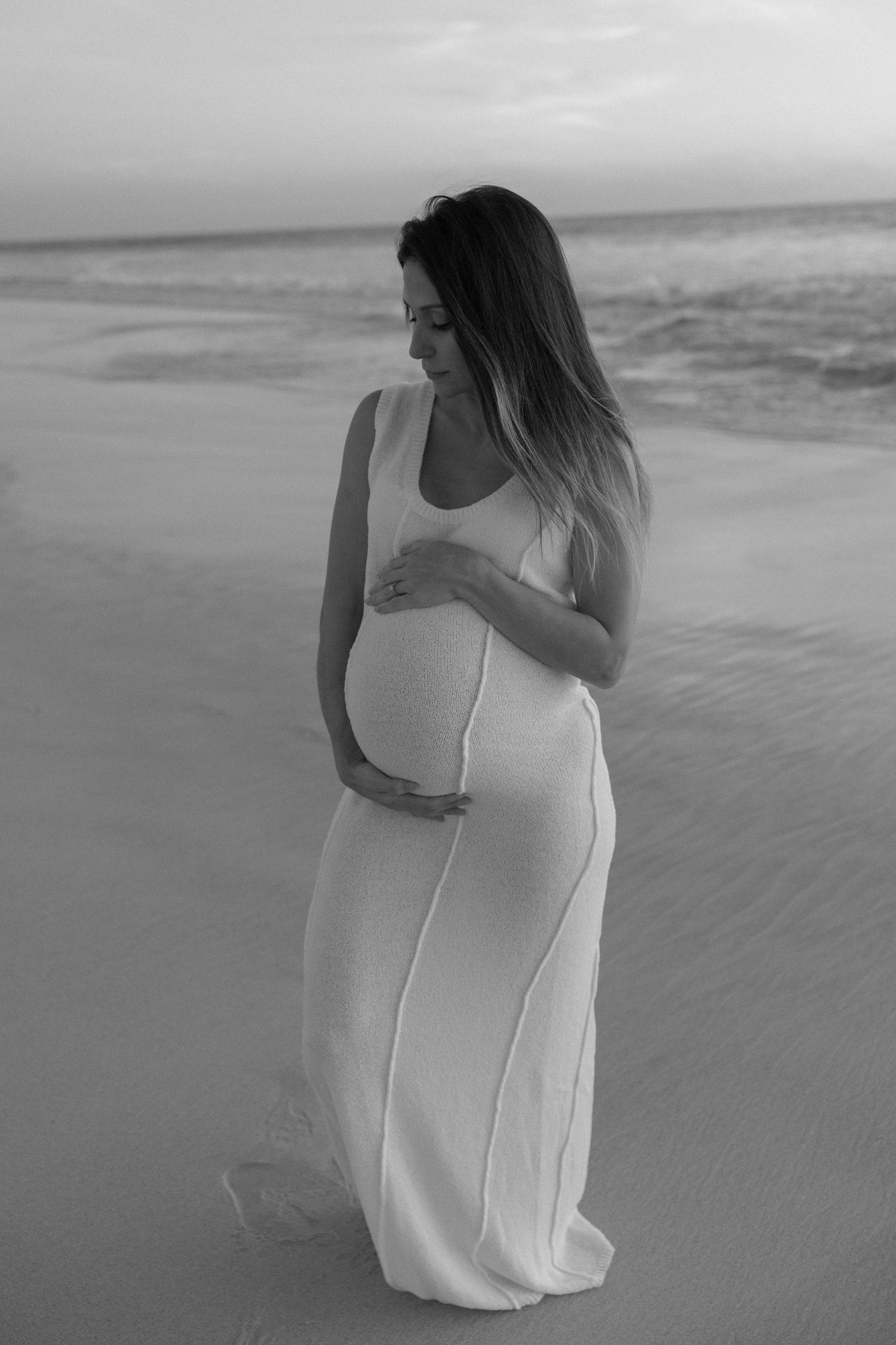 Pregnant woman in white dress cradling baby bump on beach at sunset, black and white maternity photo.