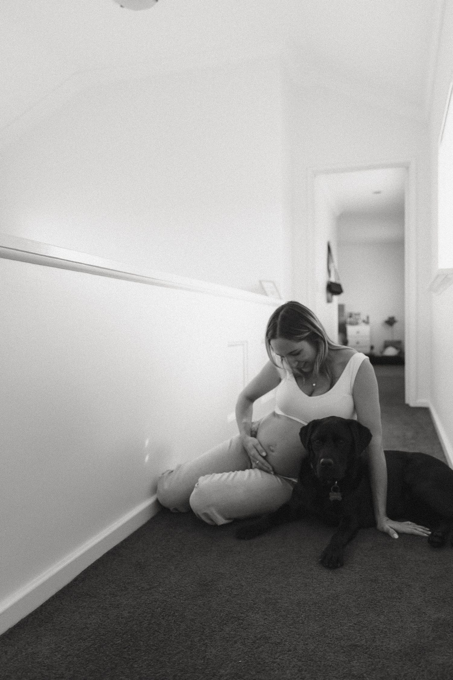 Pregnant woman sits on hallway floor with black Labrador dog in black and white maternity photo.