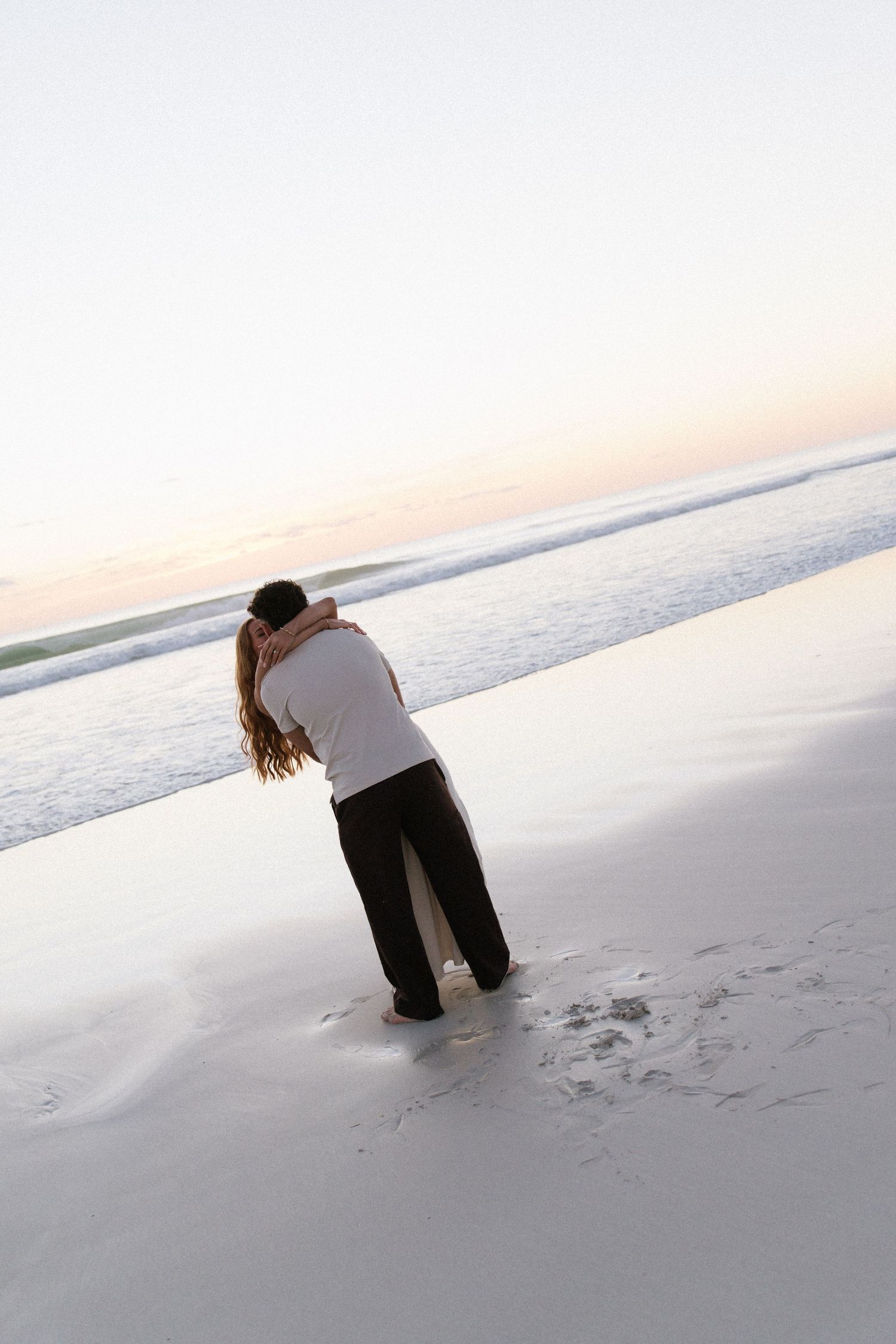 Romantic couple embracing on serene beach at dusk with gentle ocean waves behind them.