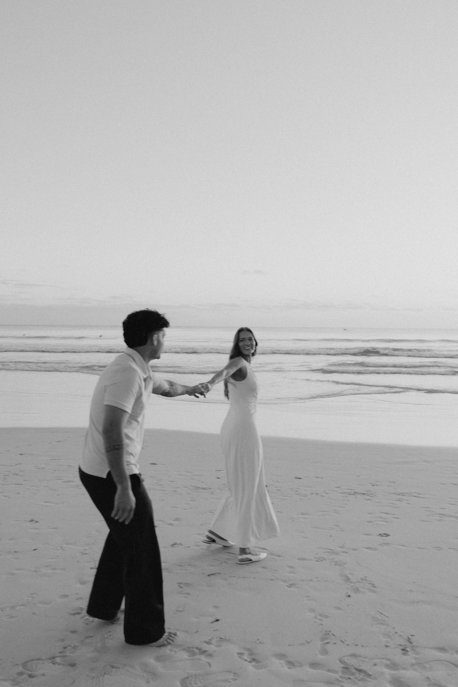 Black and white photo of a couple holding hands on a beach at sunset, woman in a flowing white dress.