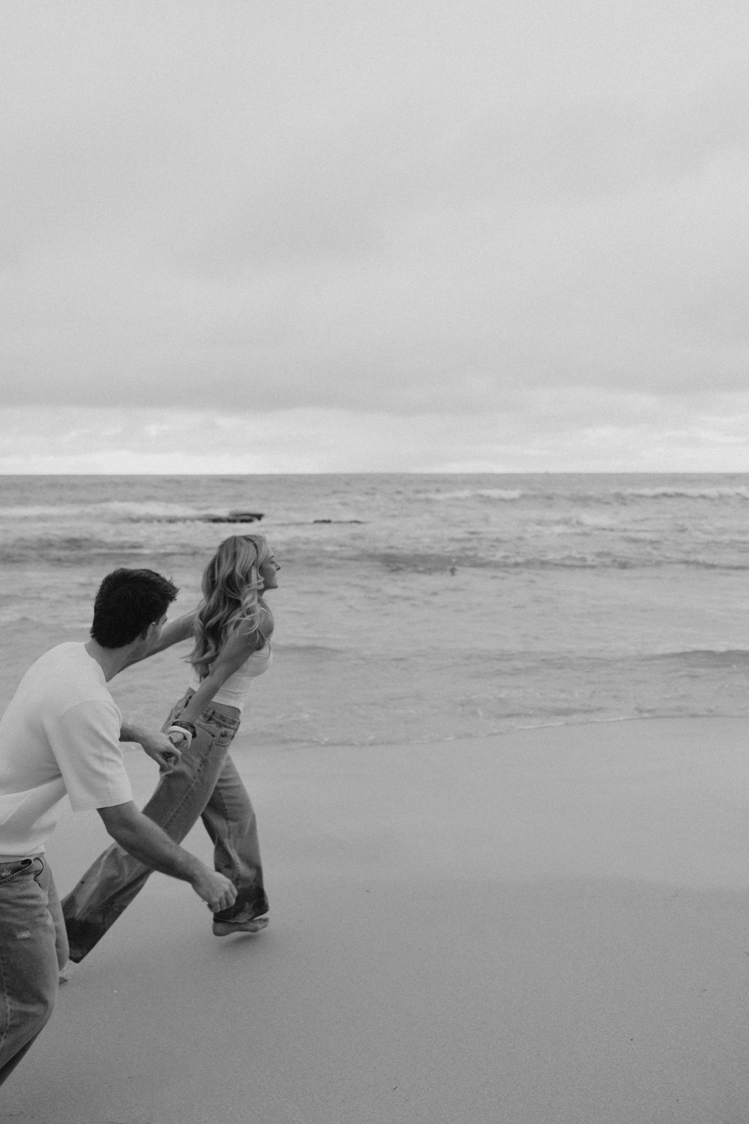 Black and white photo of couple playfully running together along a sandy beach with ocean waves.