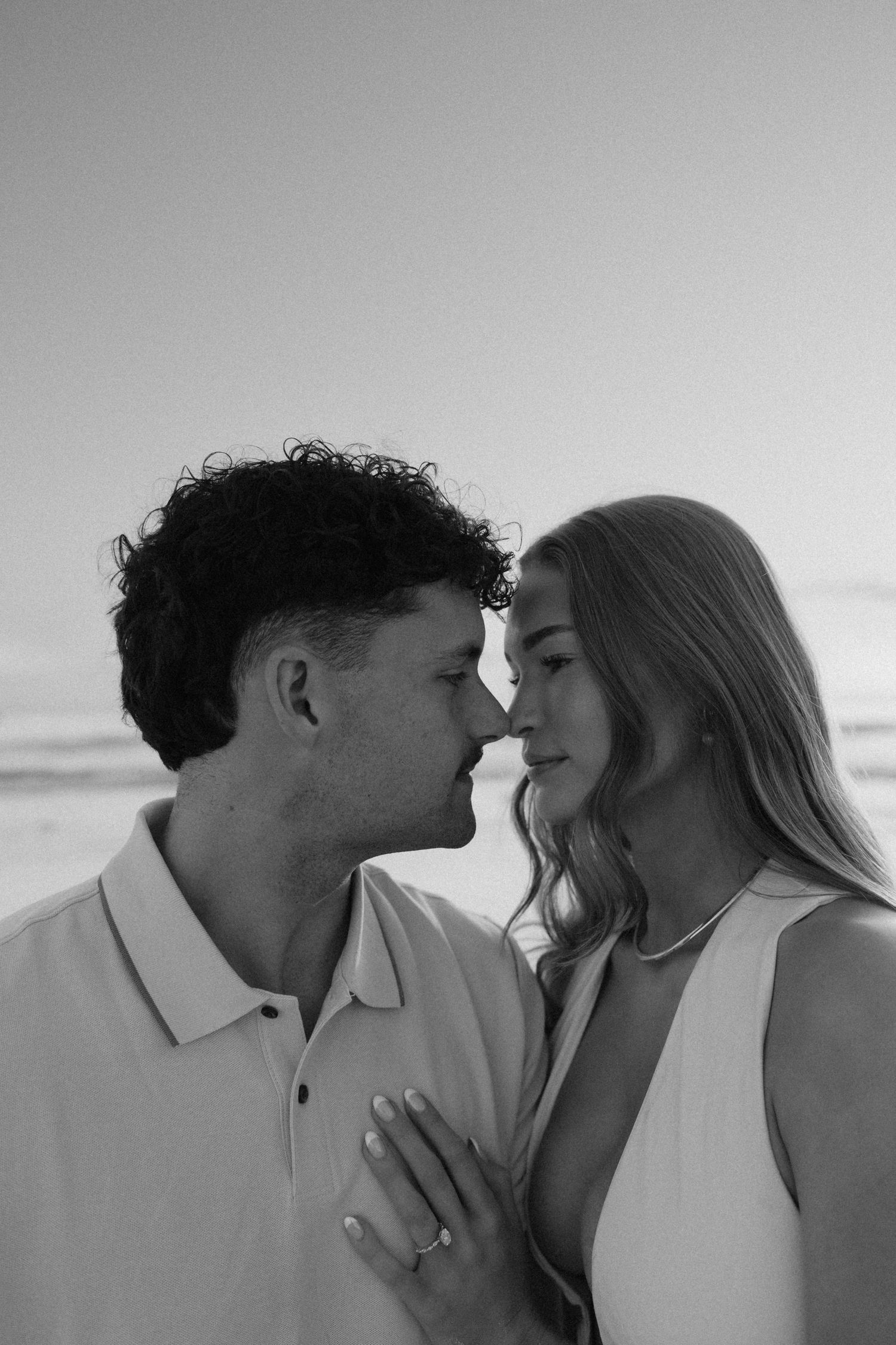 Black and white close-up of couple nearly kissing on beach, woman displaying engagement ring.