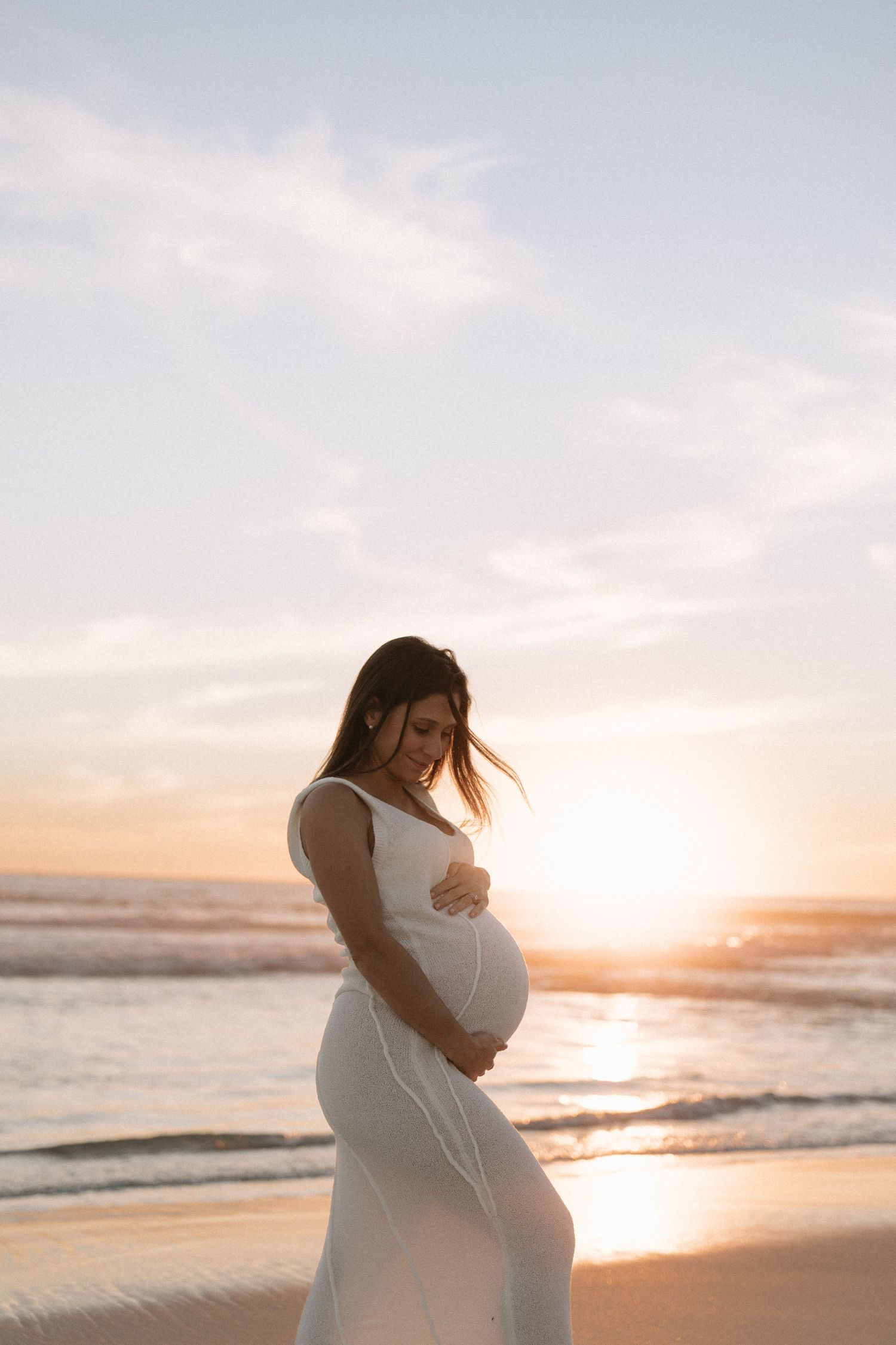 A pregnant woman in a white dress holds her belly while standing on a beach at golden sunset.