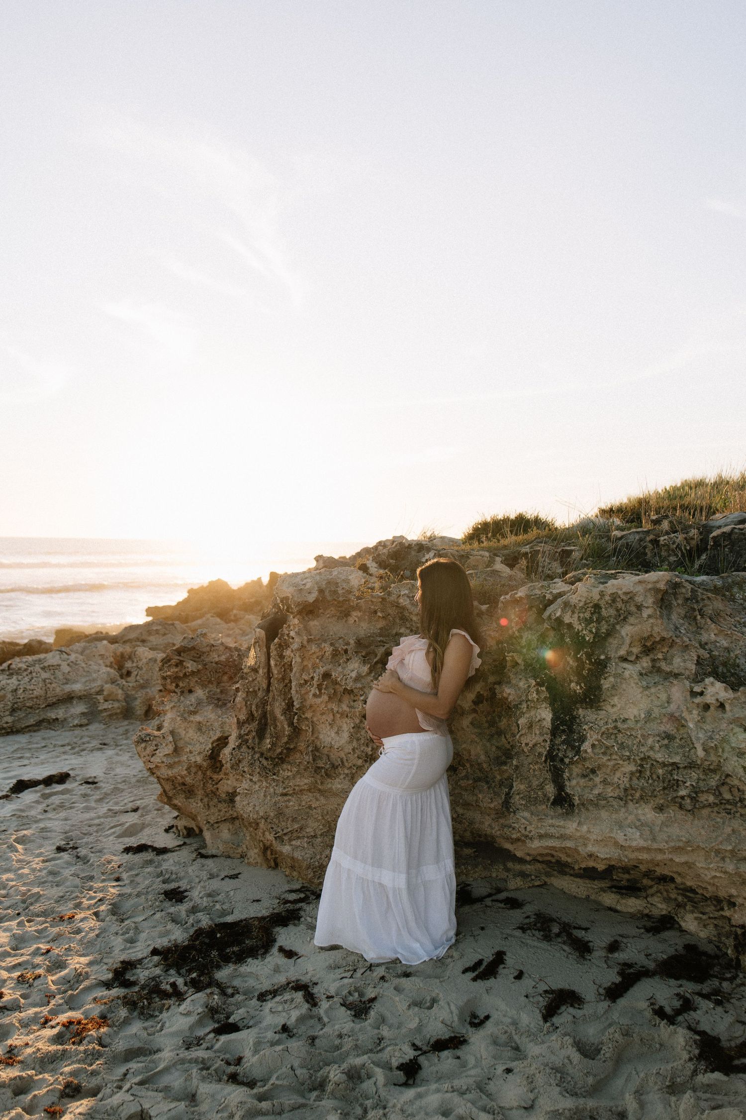 Pregnant woman in white dress standing near rocky cliffs on beach at golden hour.