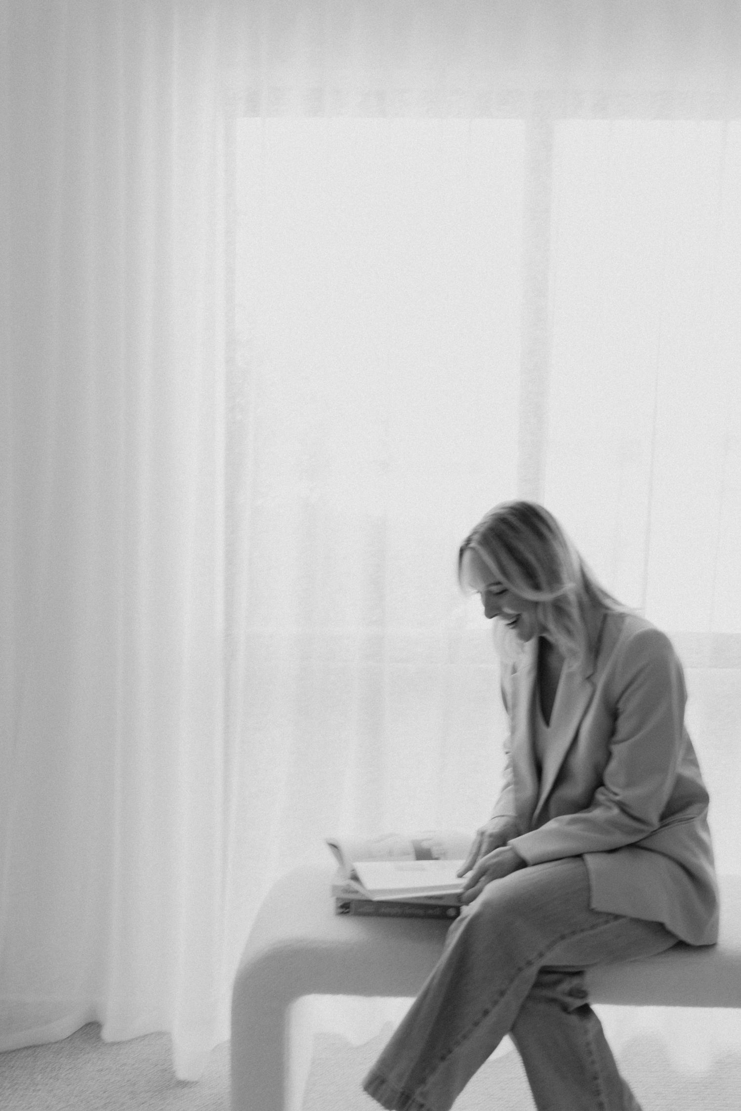 Black and white portrait of woman in blazer reading book while seated on bench by sheer curtains.