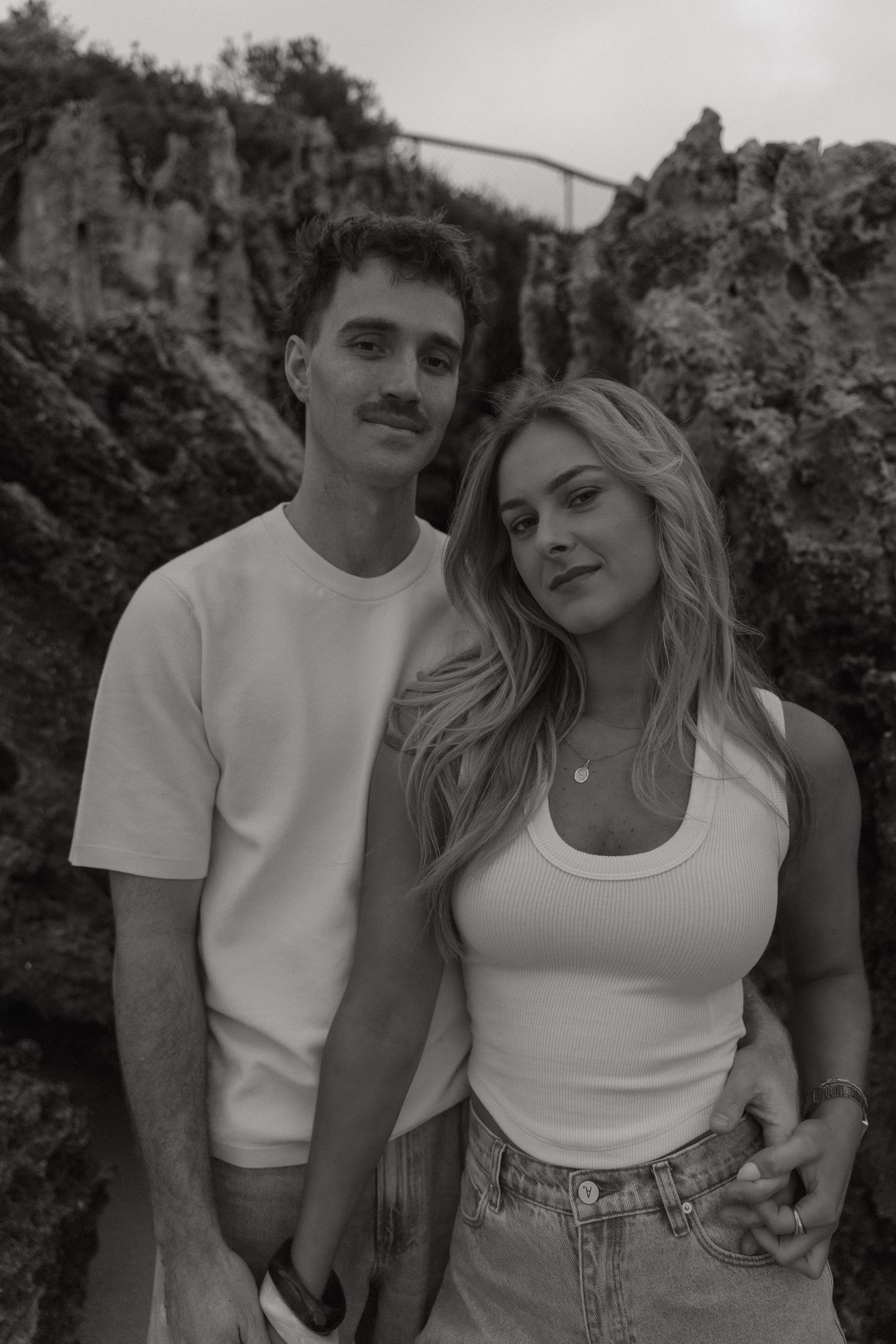 Black and white portrait of a couple standing together in front of rocky cliffs at the beach.
