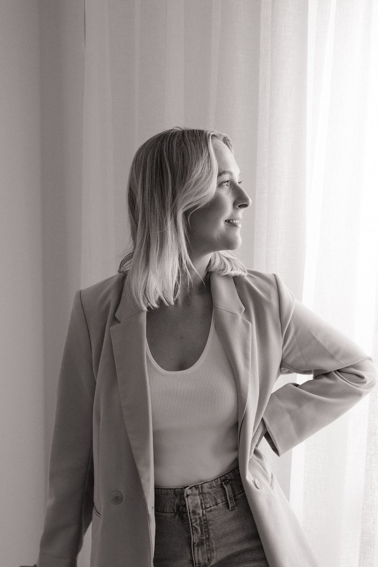 Black and white portrait of a blonde woman in a blazer looking out a window.