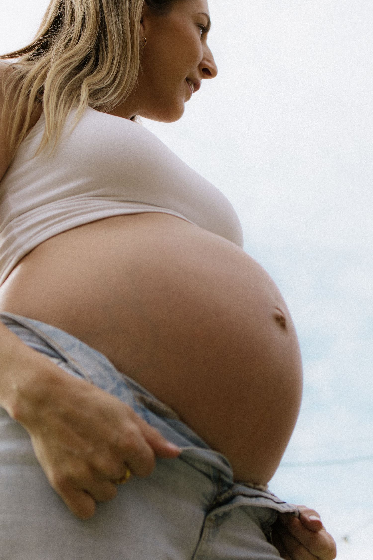 Pregnant woman in white crop top and jeans cradling her bare baby bump outdoors.