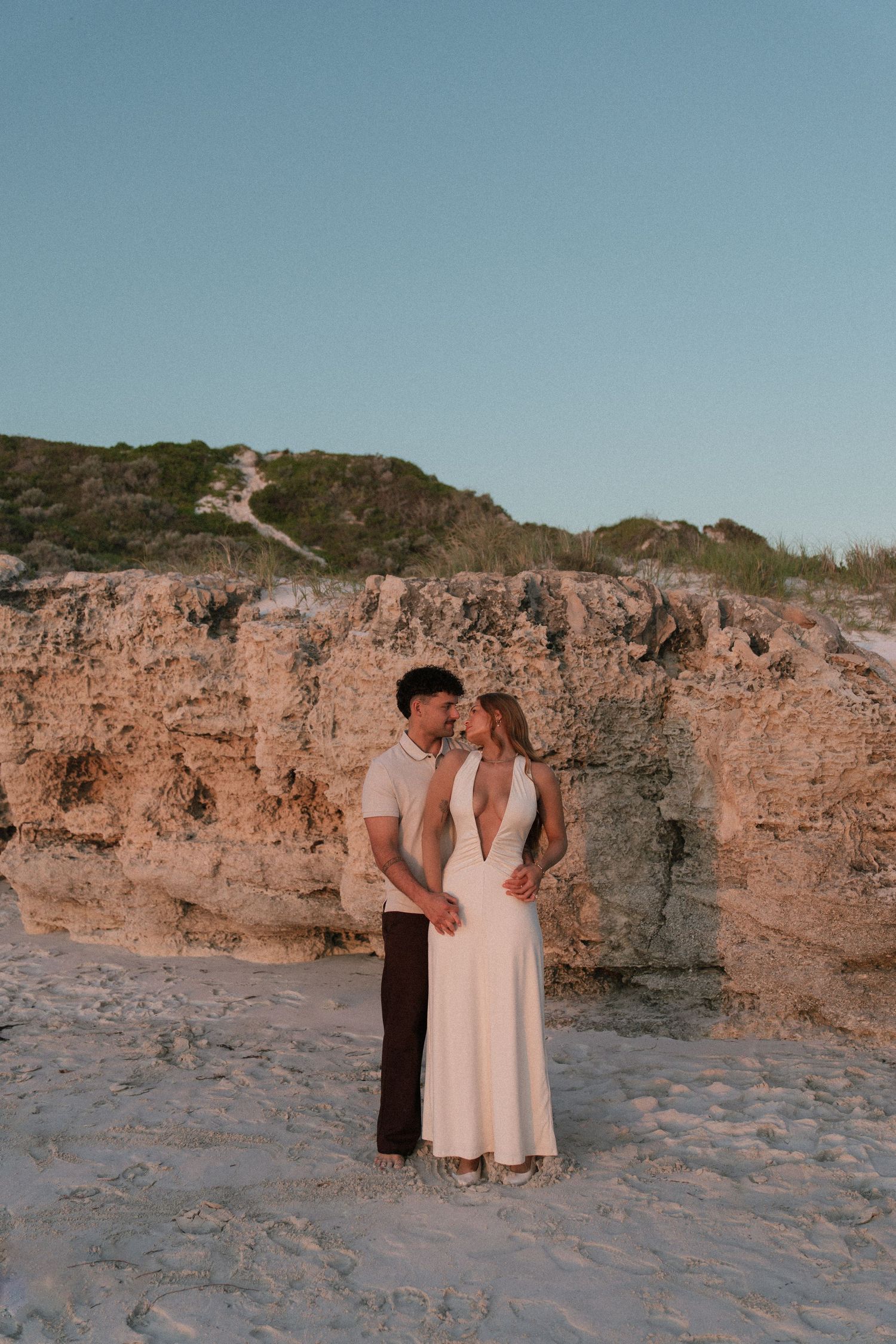 Couple sharing a kiss near rustic rock formations on a sandy beach at dusk.