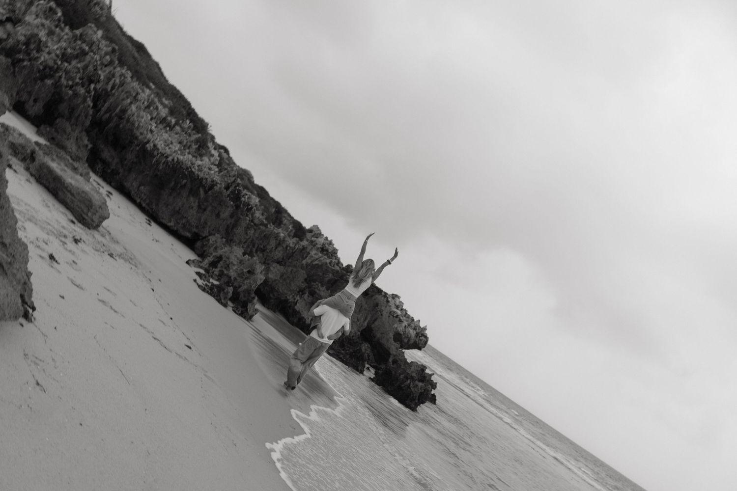 Black and white image of a person jumping joyfully on a sandy beach near rocky cliffs.