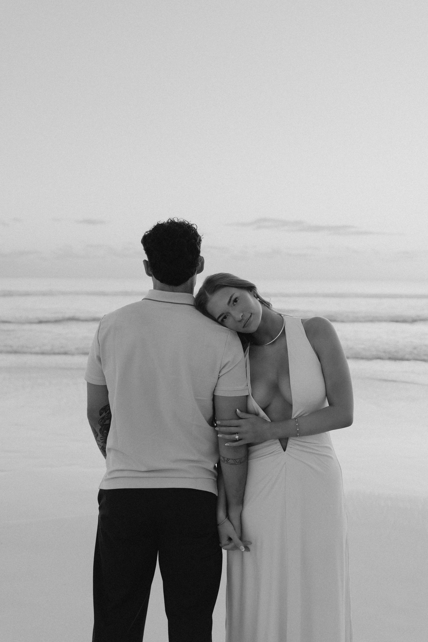 Black and white photo of a woman in white dress leaning on her partners shoulder at the beach.