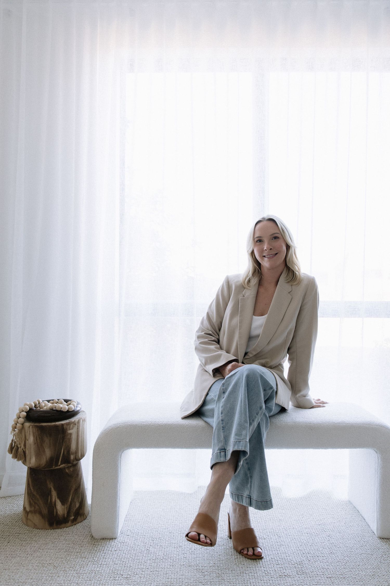 Smiling blonde woman in beige blazer and jeans sitting on modern white bench near sheer curtains.