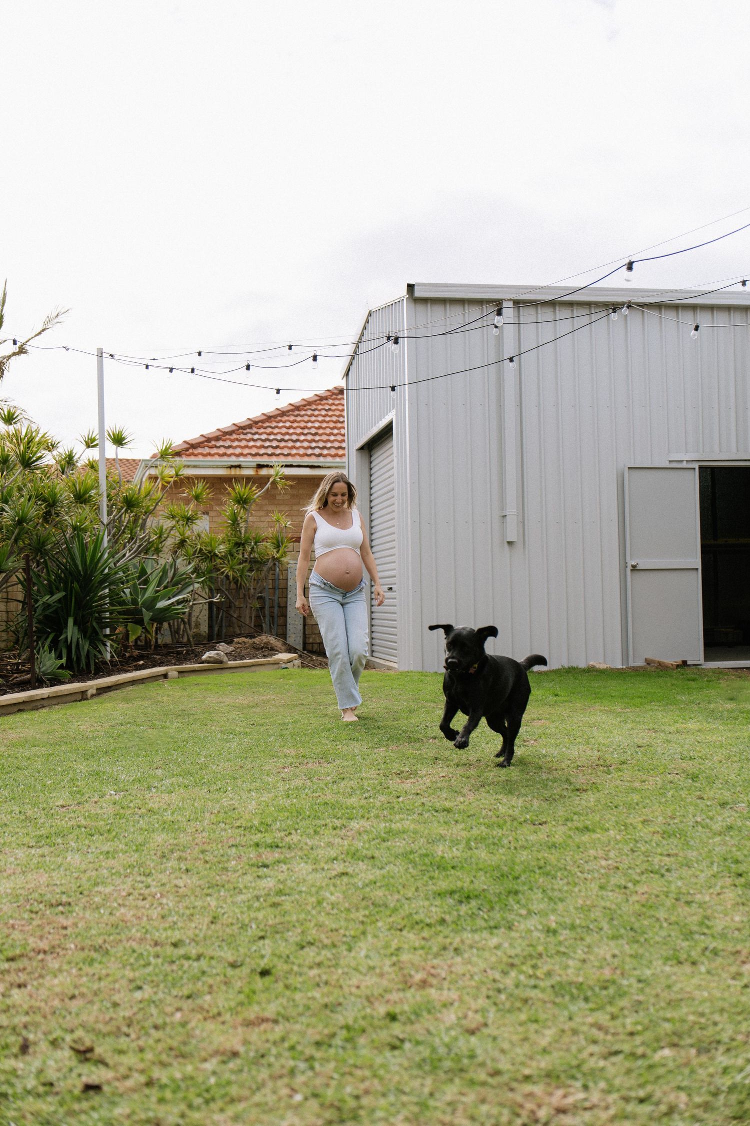 Pregnant woman in white crop top and jeans playing with black dog in backyard garden.
