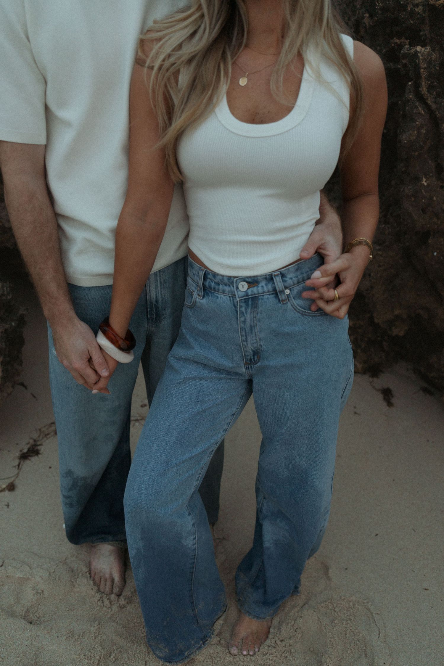 Close-up of a couple holding hands on a beach, woman wearing a white tank top and wide-leg jeans.