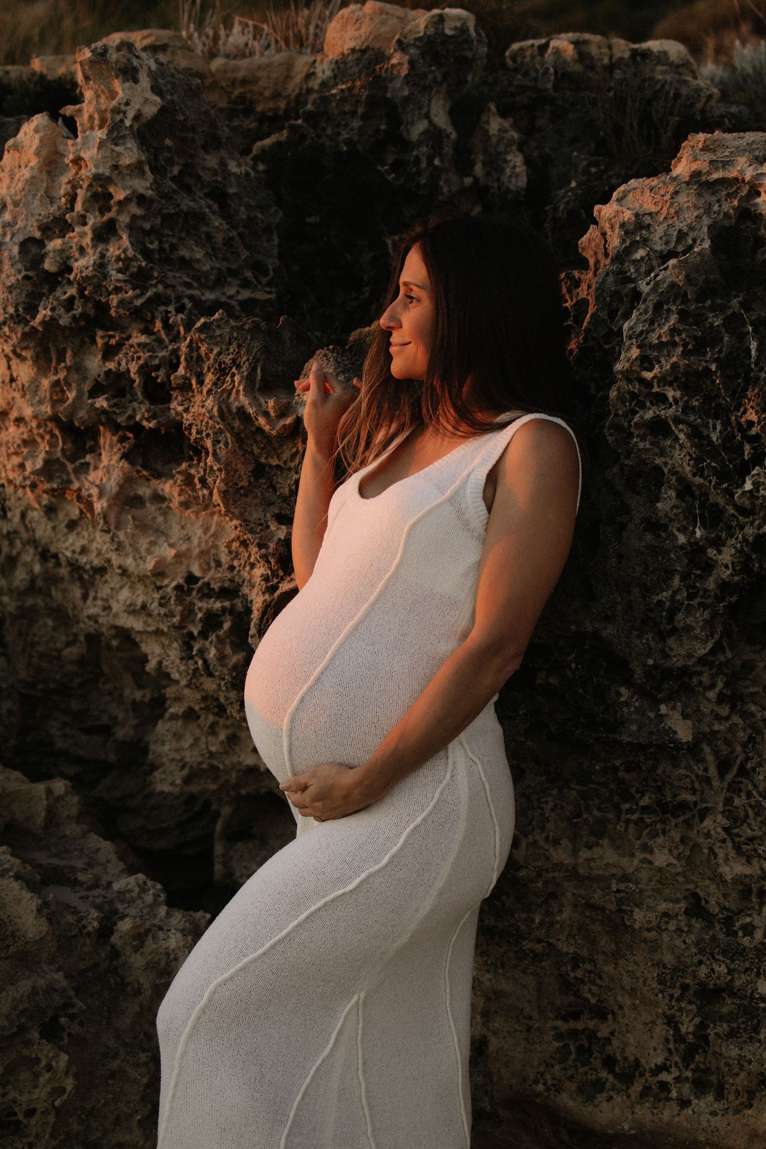 Pregnant woman in white dress cradling belly against rocky wall in golden sunset light.