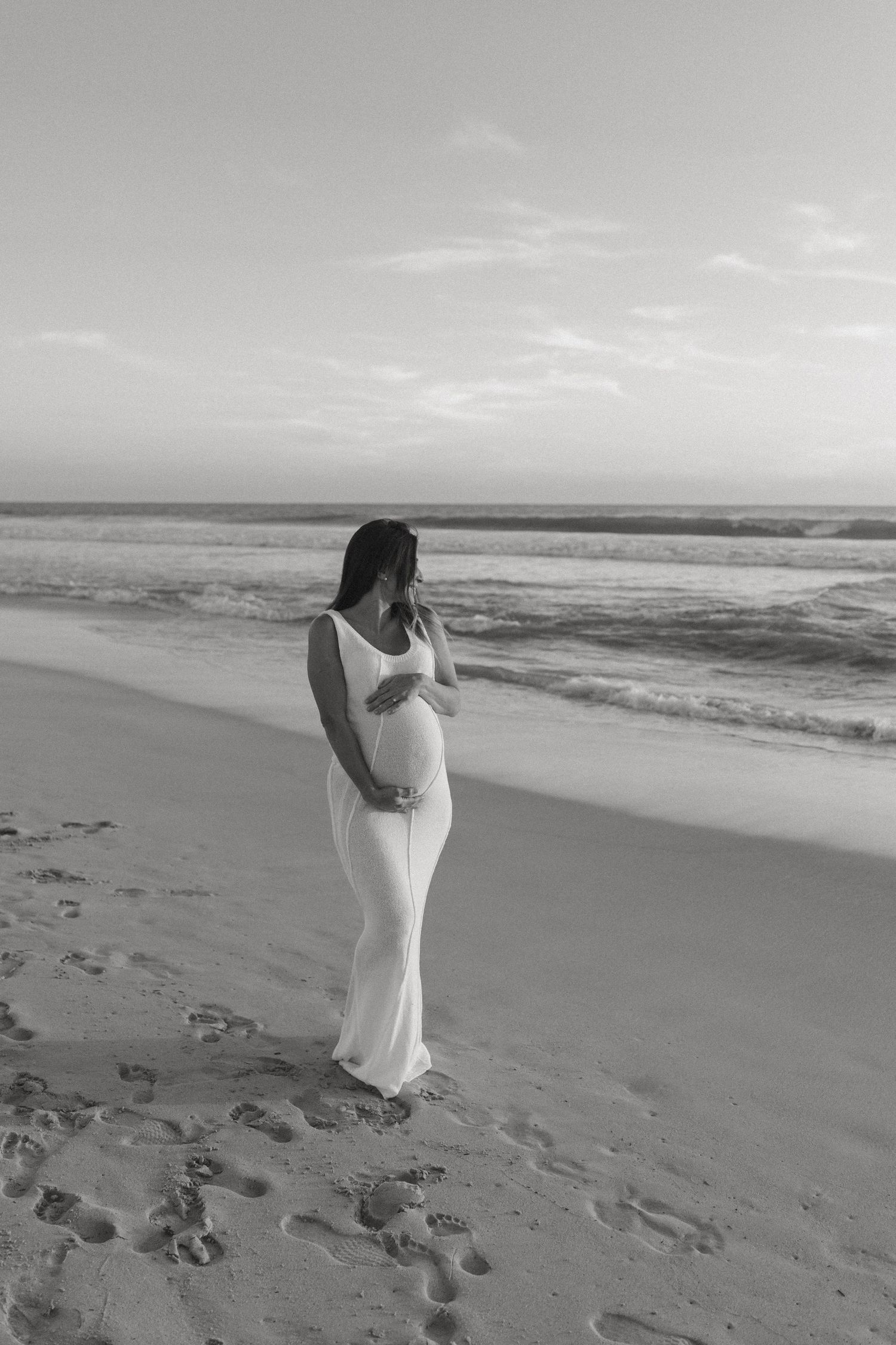 A pregnant woman in a white dress stands on a beach holding her belly, captured in black and white.