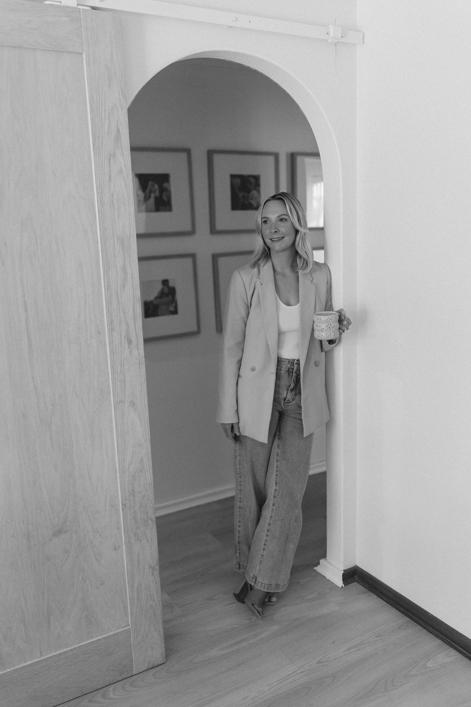 Black and white photo of smiling blonde woman in blazer holding mug standing in arched doorway.