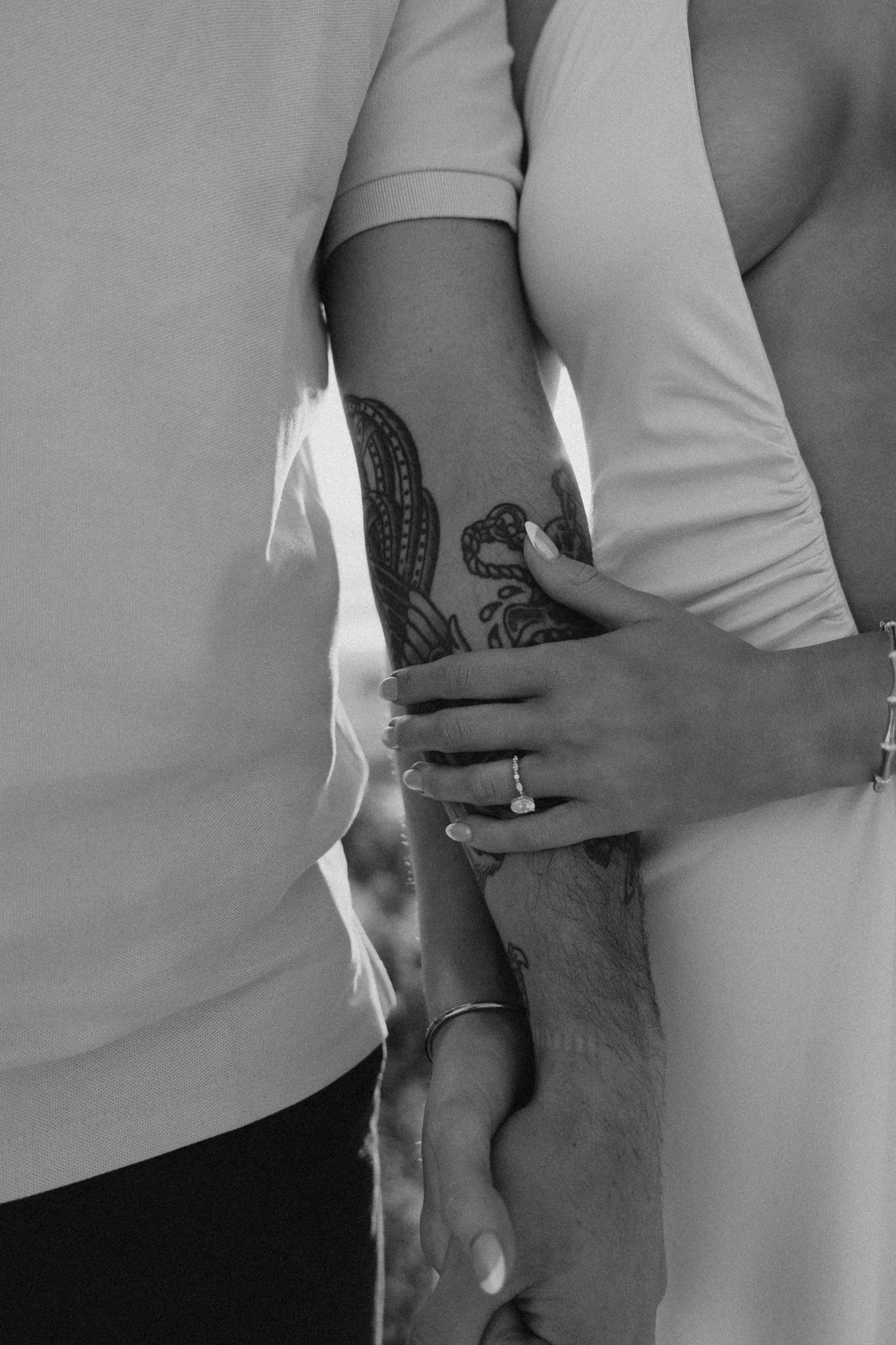 Black and white close-up of a couple embracing, man with tattoo on forearm, woman wearing engagement ring.