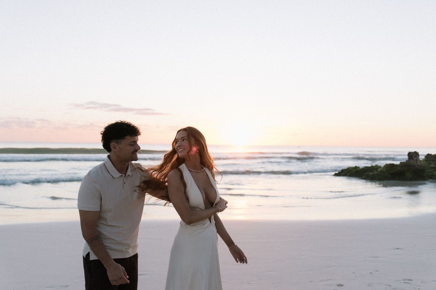 Romantic couple laughing together on a beach at sunset, woman in white dress, golden hour light.