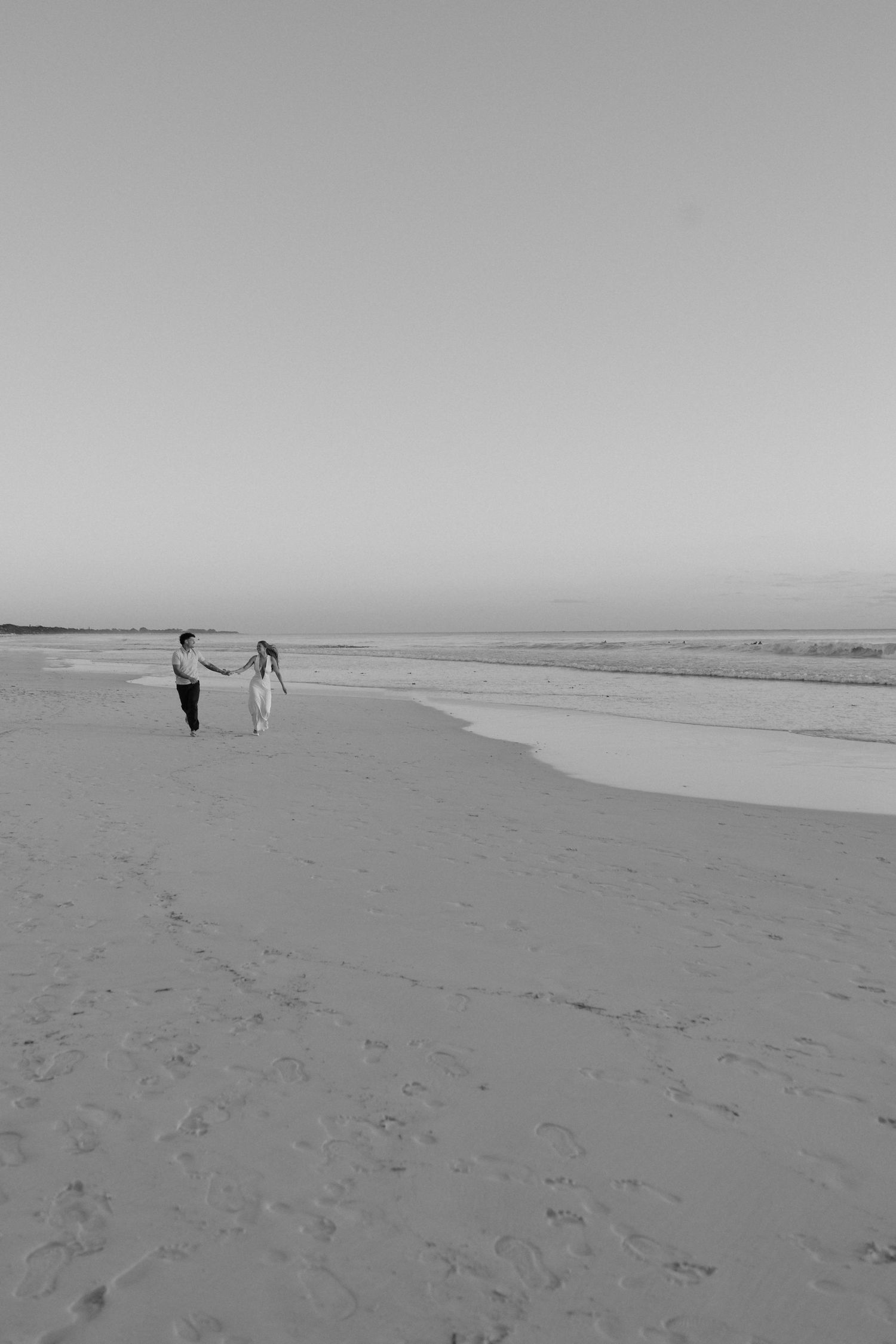 Black and white wide shot of couple holding hands walking on vast empty beach at dusk.