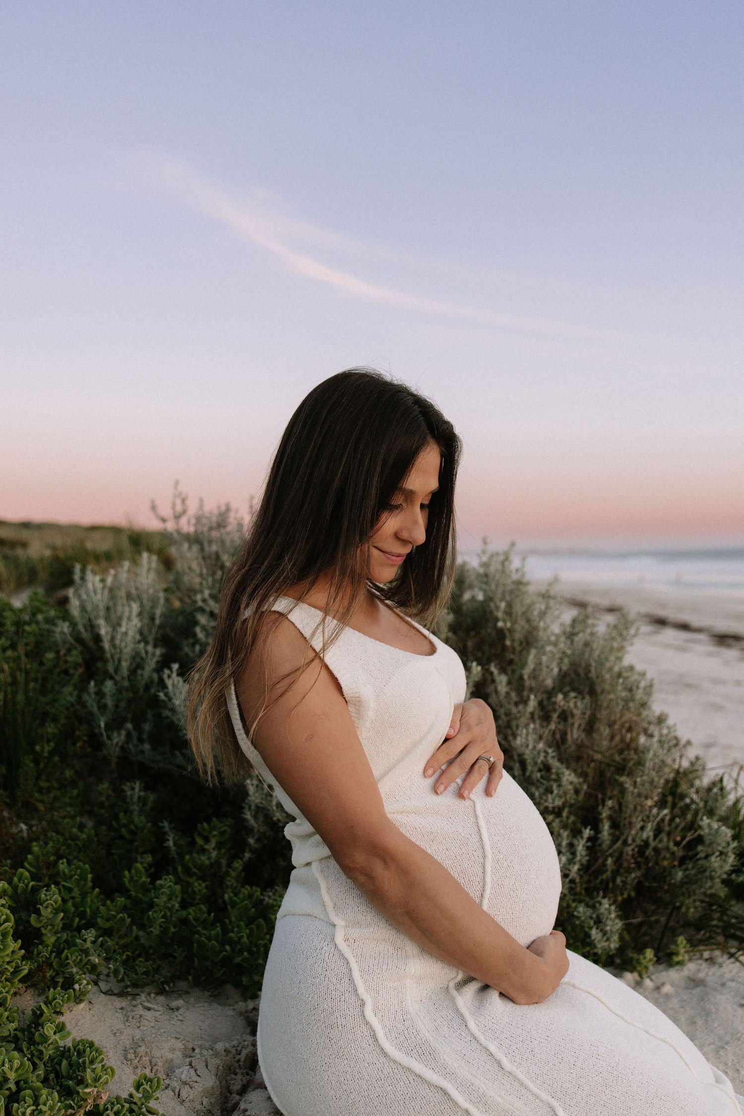 Pregnant woman in white dress sitting among beach dunes at pink sunset, holding belly.