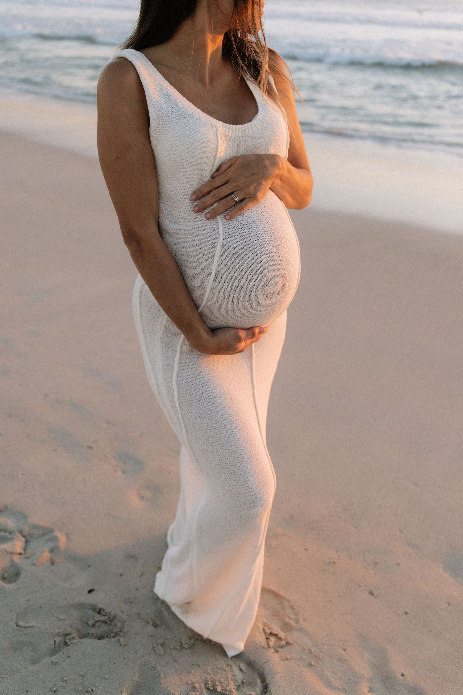 Pregnant woman in white knit dress cradling baby bump on sandy beach at sunset.