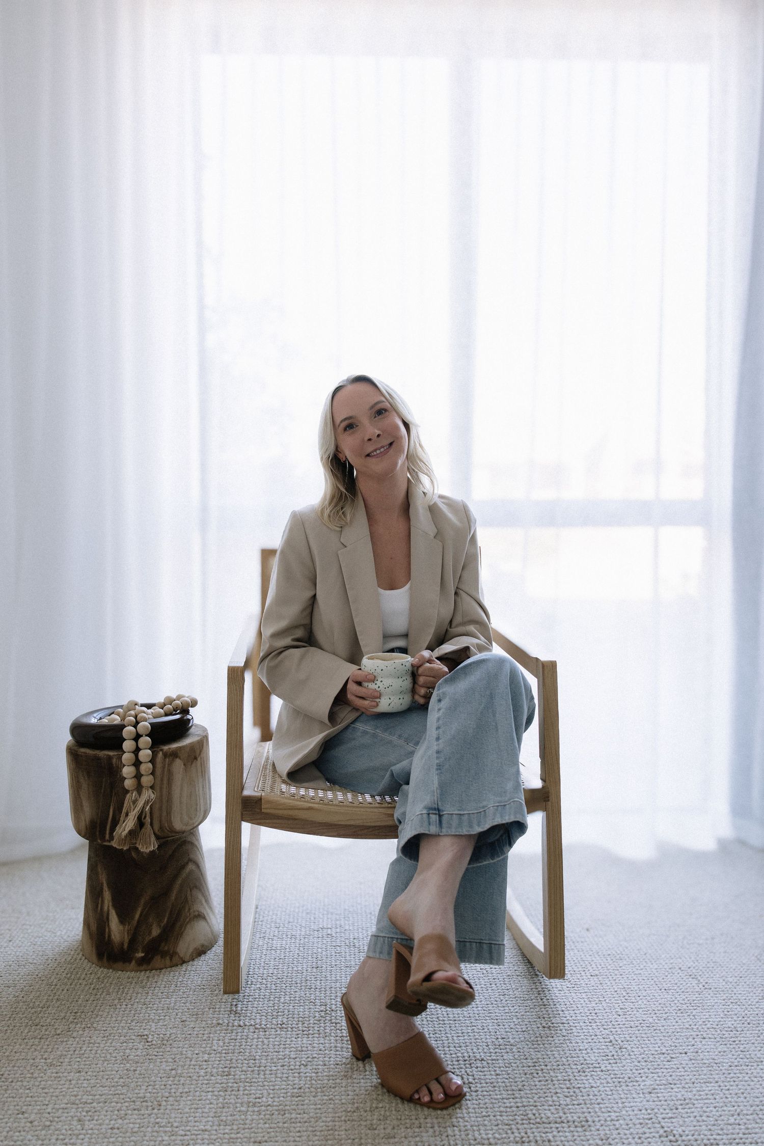 Smiling blonde woman in beige blazer holding mug and book while relaxing in wooden chair by window.