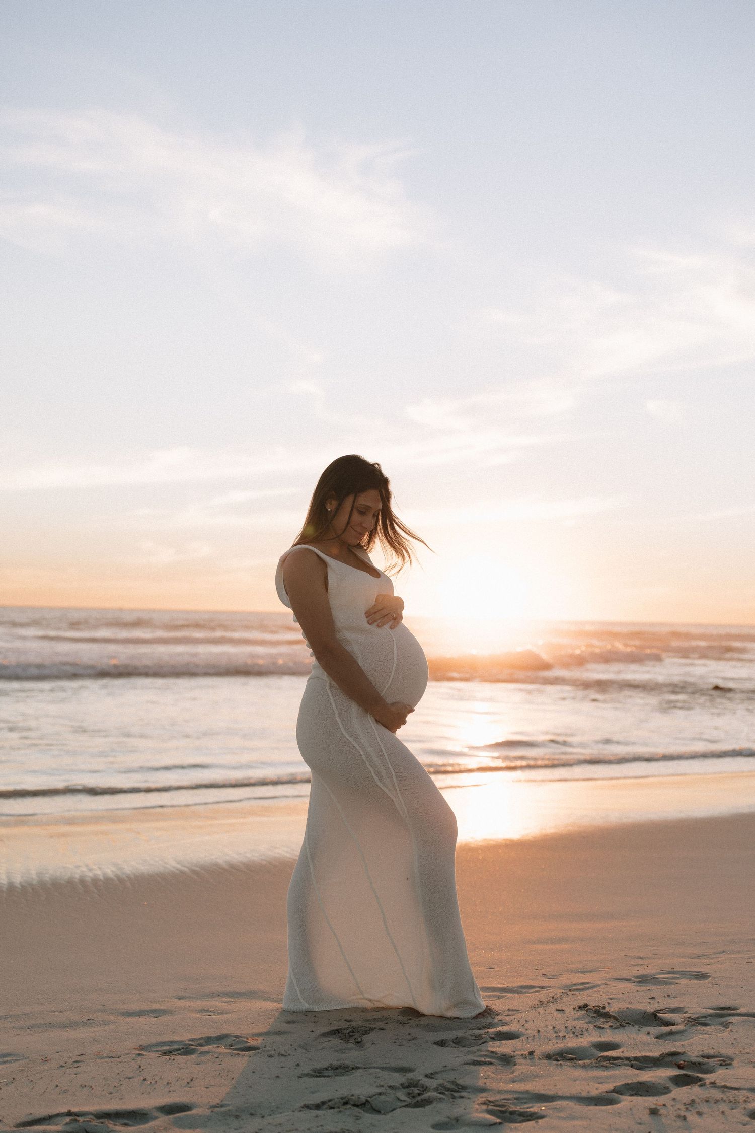 A pregnant woman in a flowing white dress cradles her bump on a sandy beach at sunset.