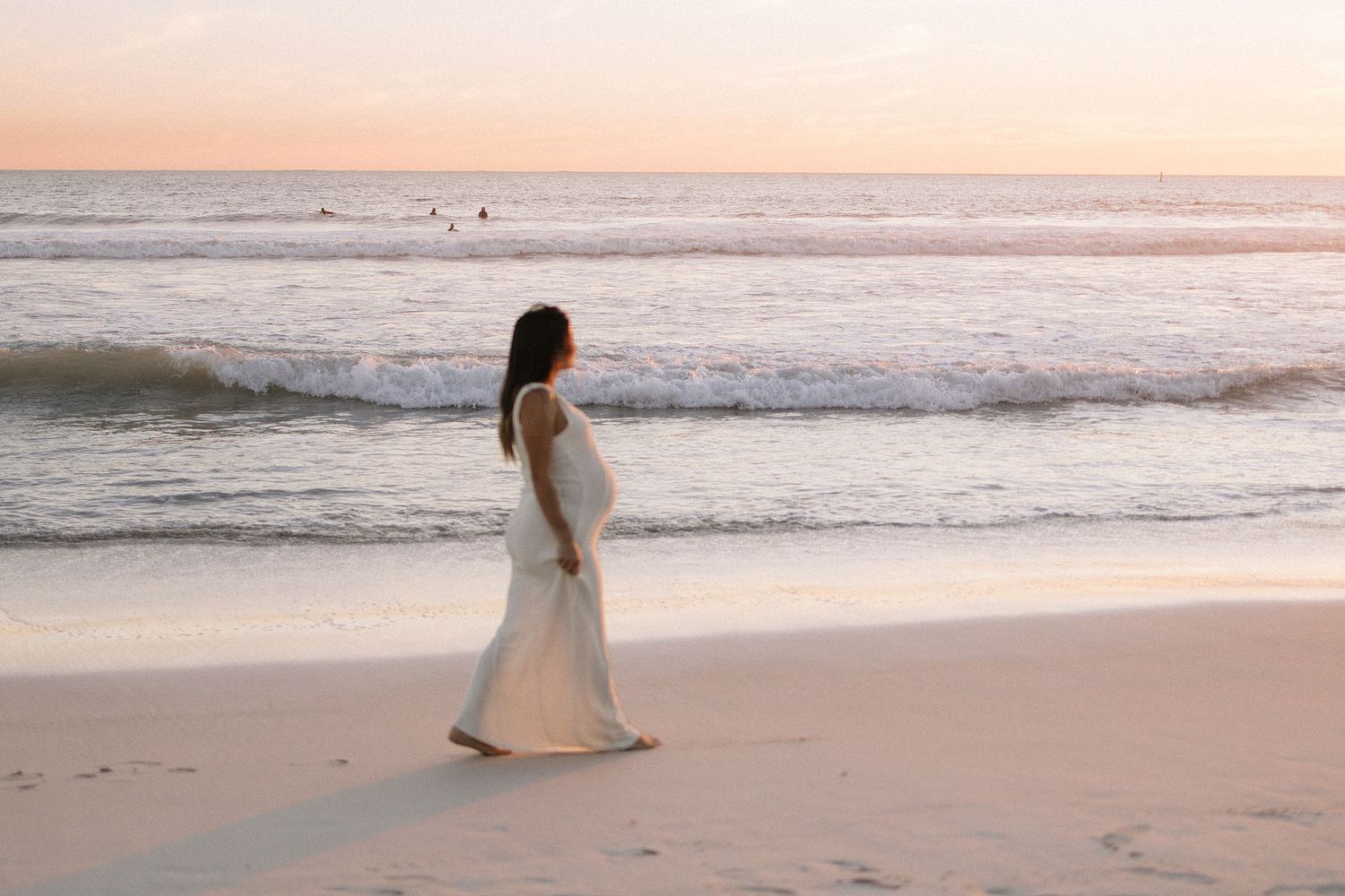 Pregnant woman in white maxi dress standing alone on beach at pastel sunset.