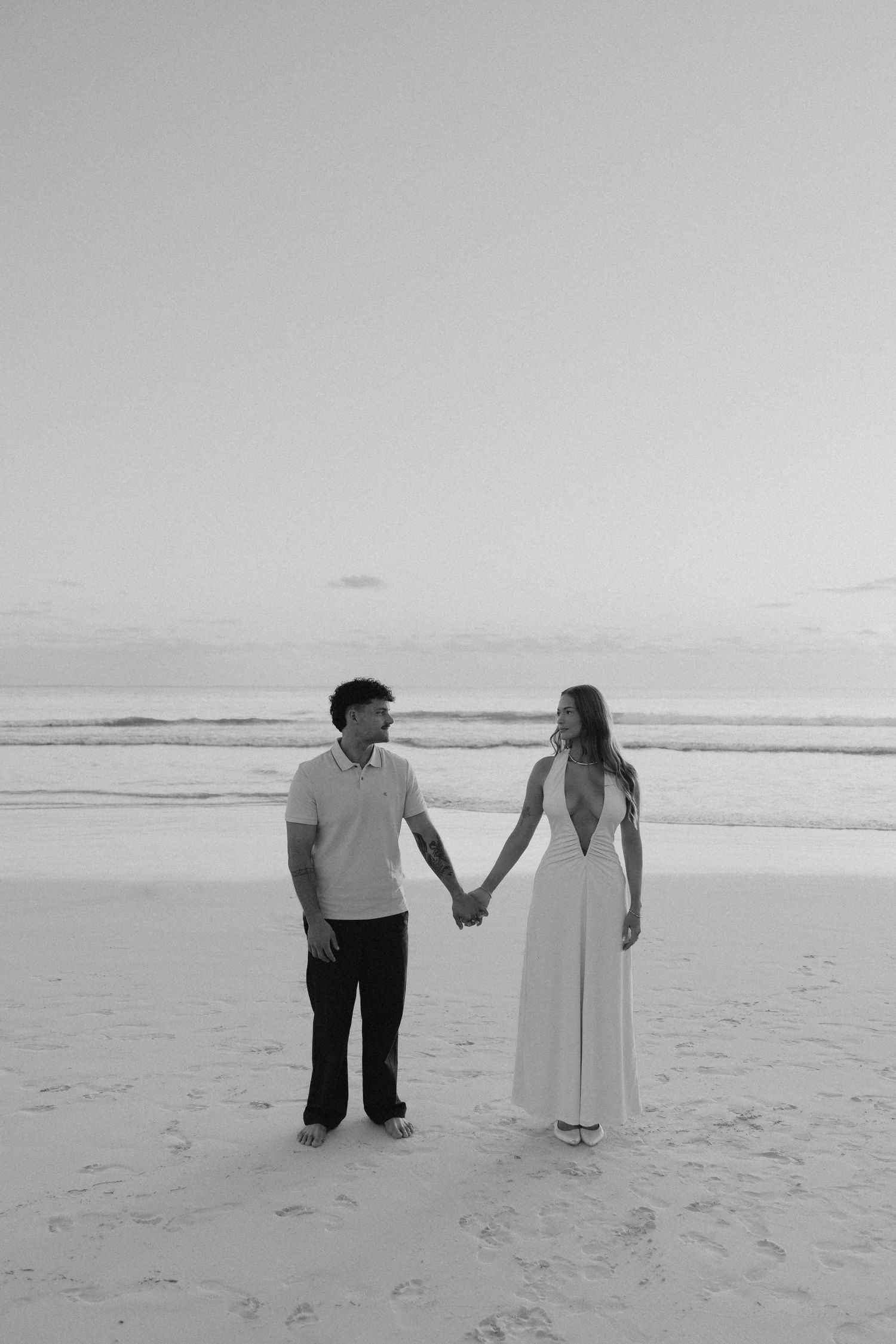 Black and white photo of a couple holding hands on a beach, woman in white backless gown, ocean backdrop.