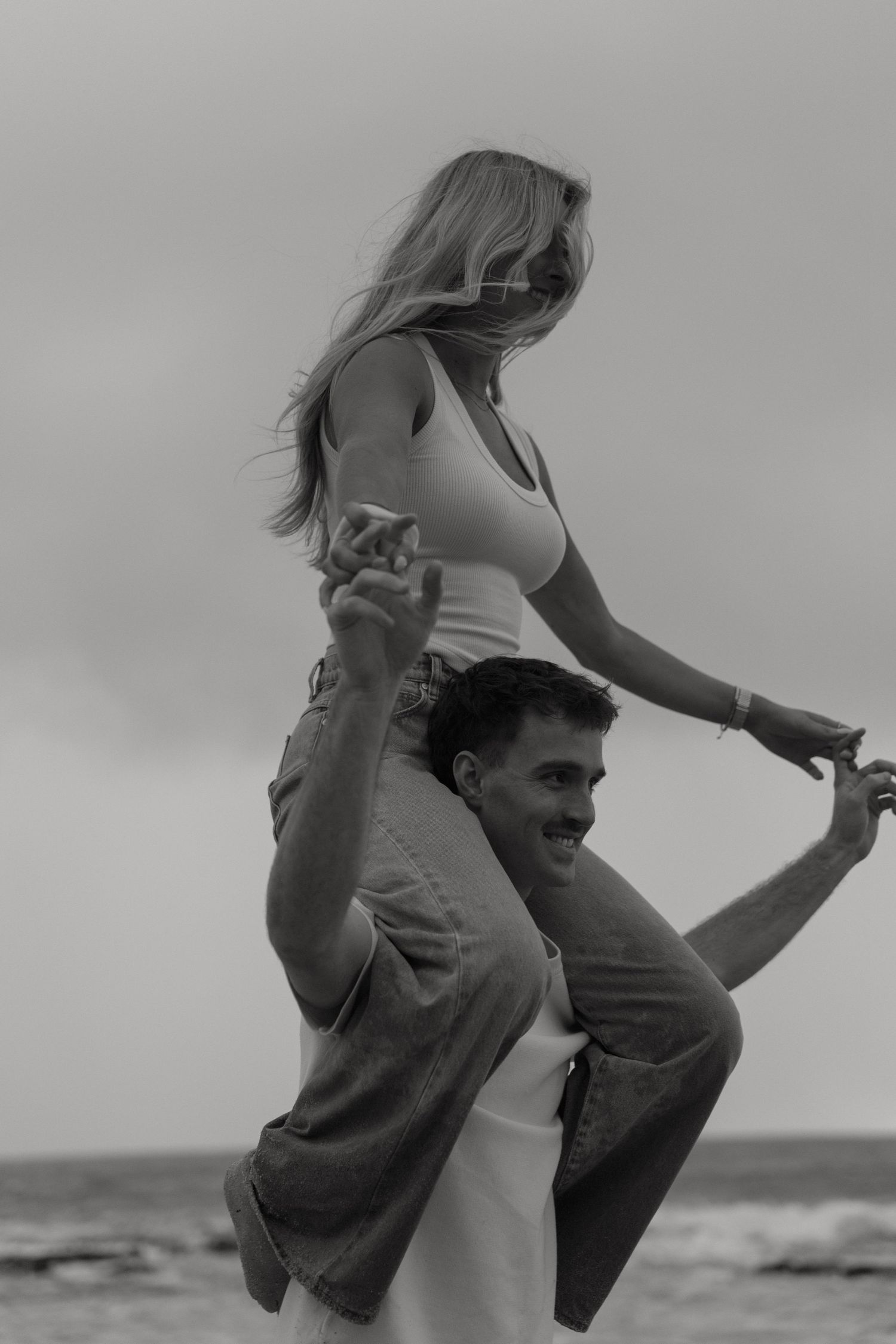 Black and white photo of a smiling man carrying a woman on his shoulders at a windy beach.