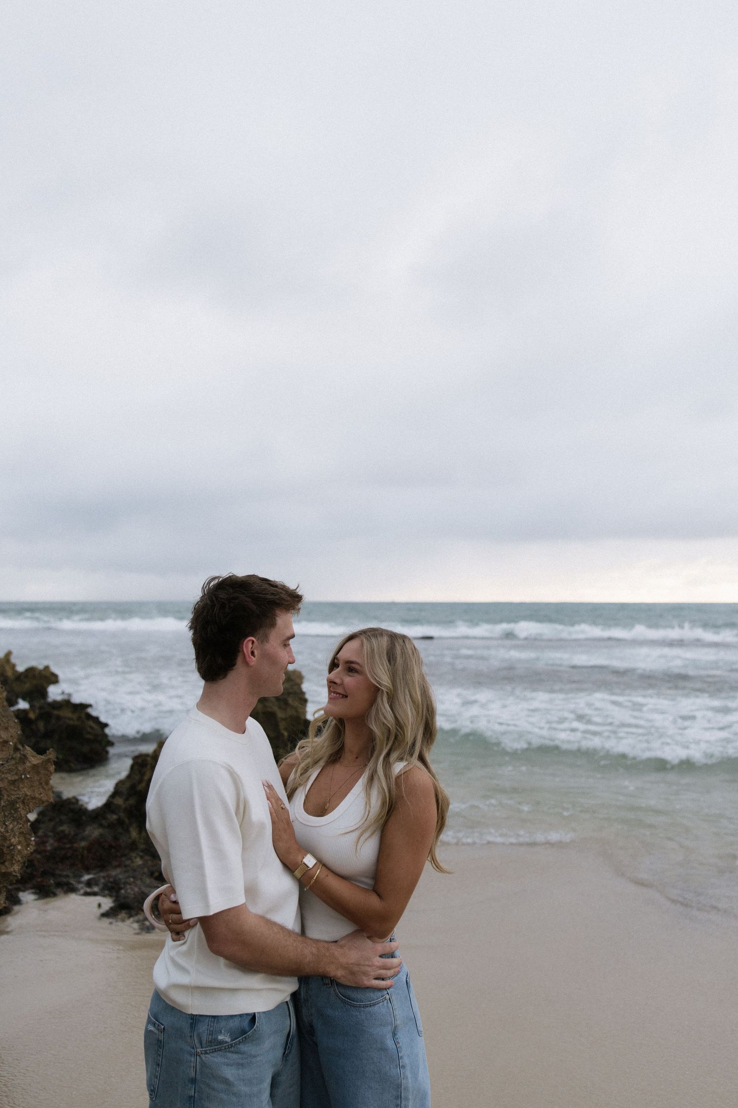 Couple embracing and looking at each other on a beach with ocean waves and rocky cliffs behind them.