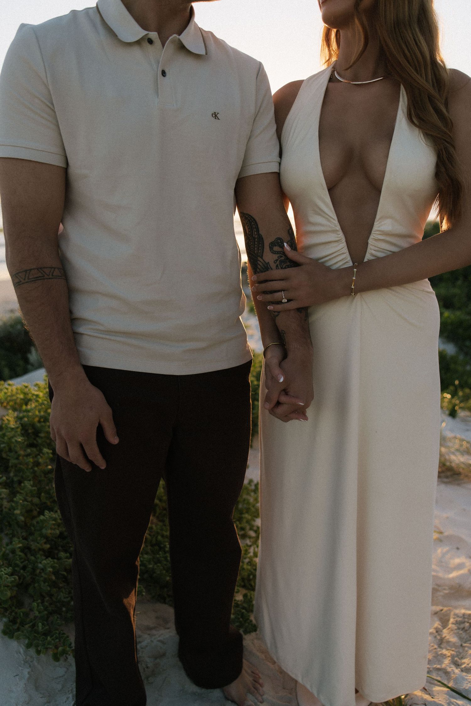 Couple holding hands at sunset, man in polo shirt and dark pants, woman in a deep-V cream halter dress.