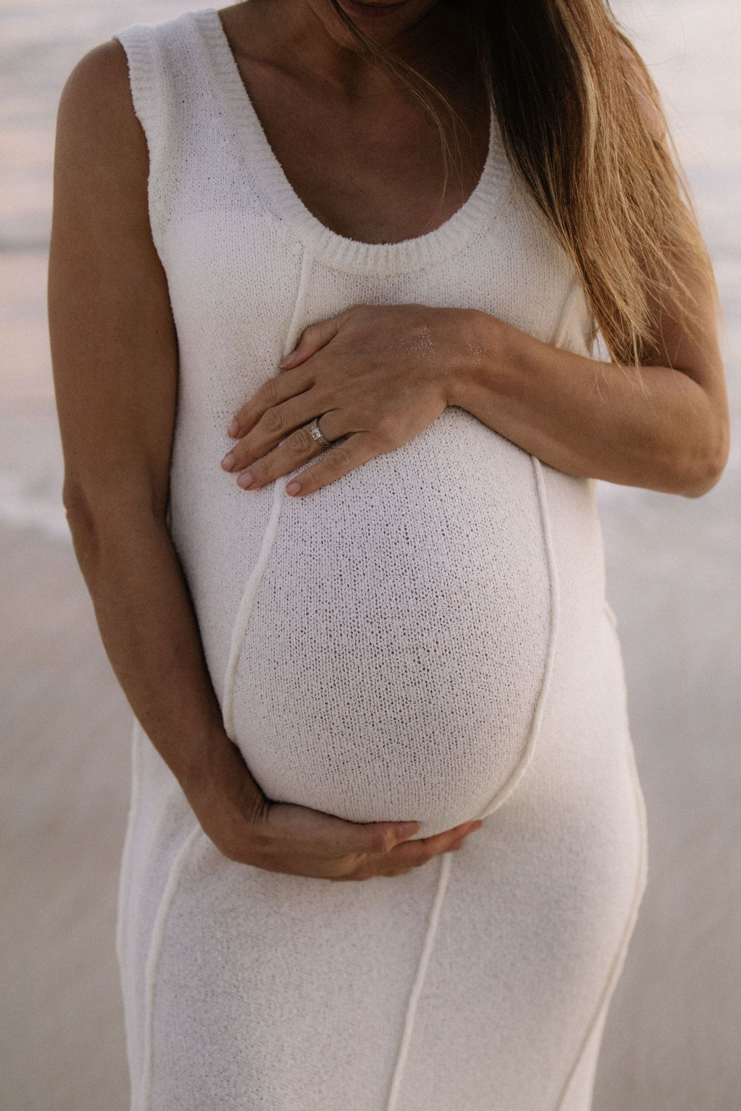 Close-up of pregnant woman in white knit dress holding baby bump.
