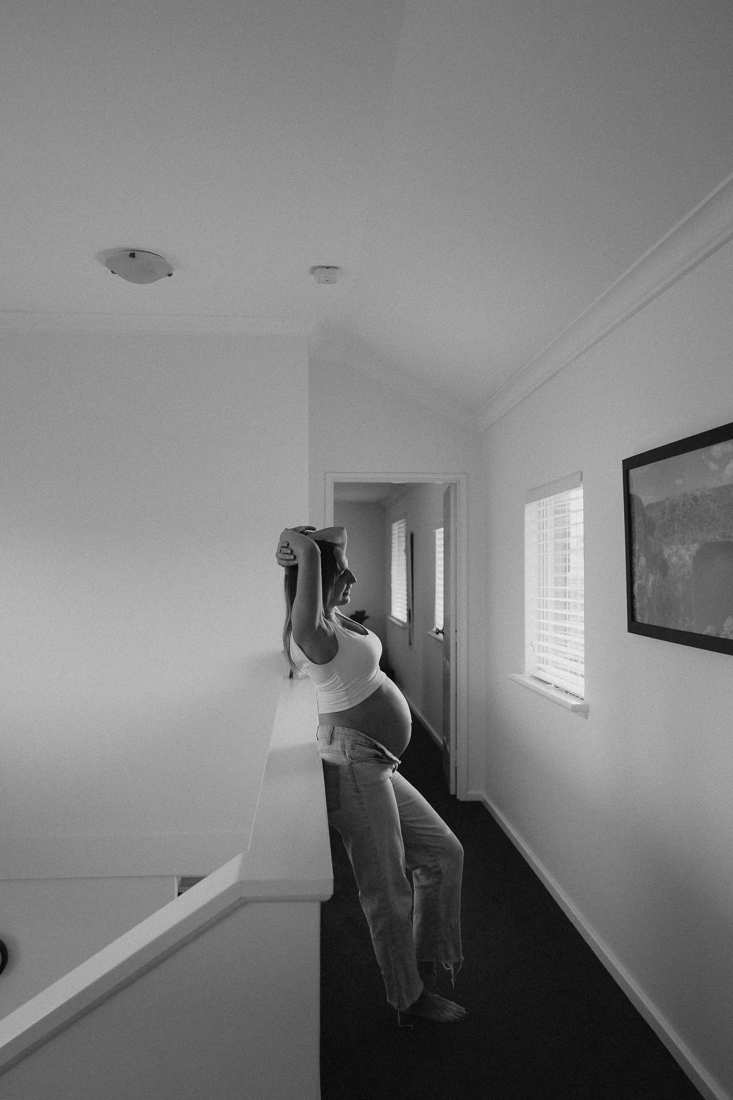 Pregnant woman in white top and jeans leans against hallway railing, black and white maternity photo.