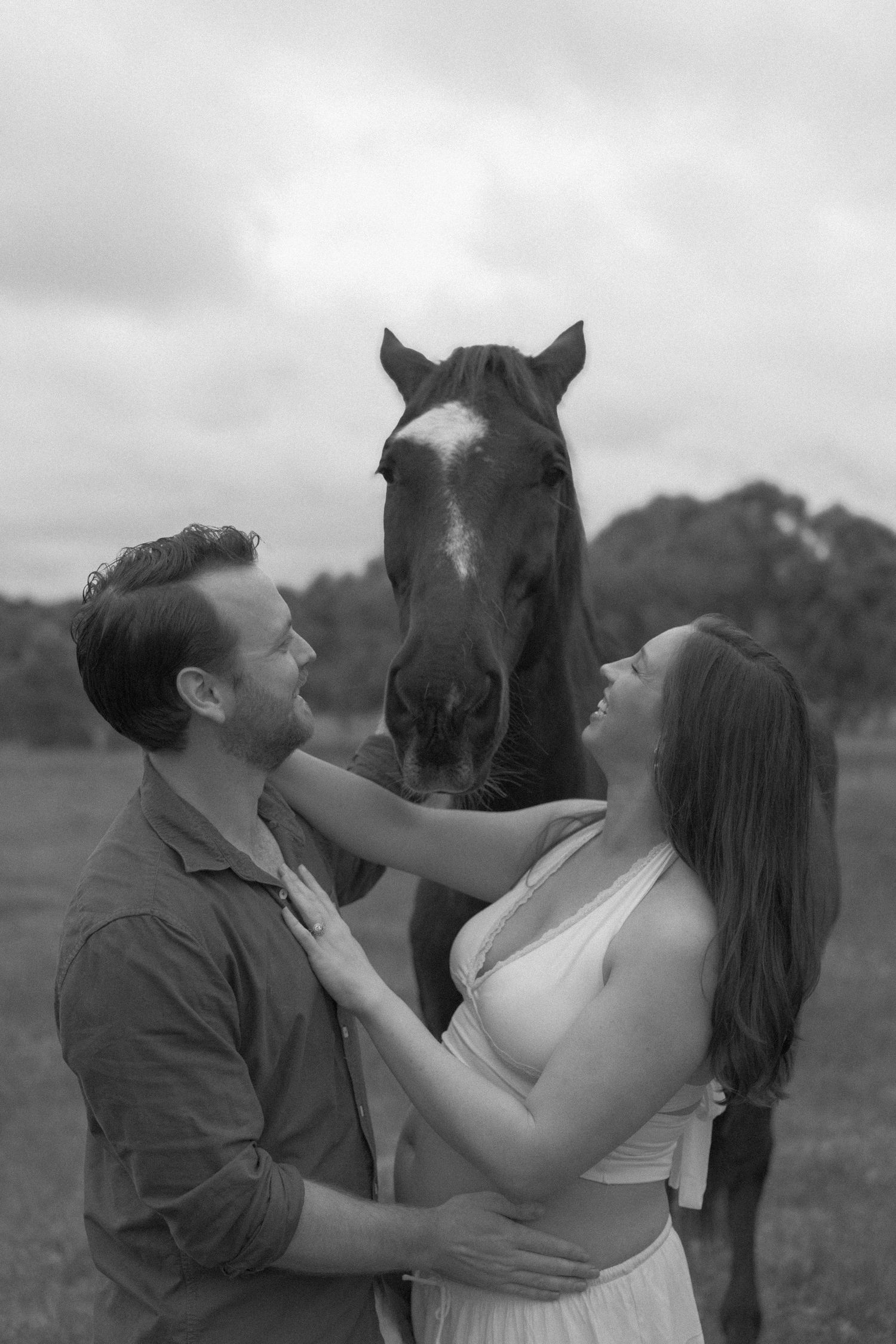 Black and white photo of couple smiling with a horse between them during outdoor maternity session.