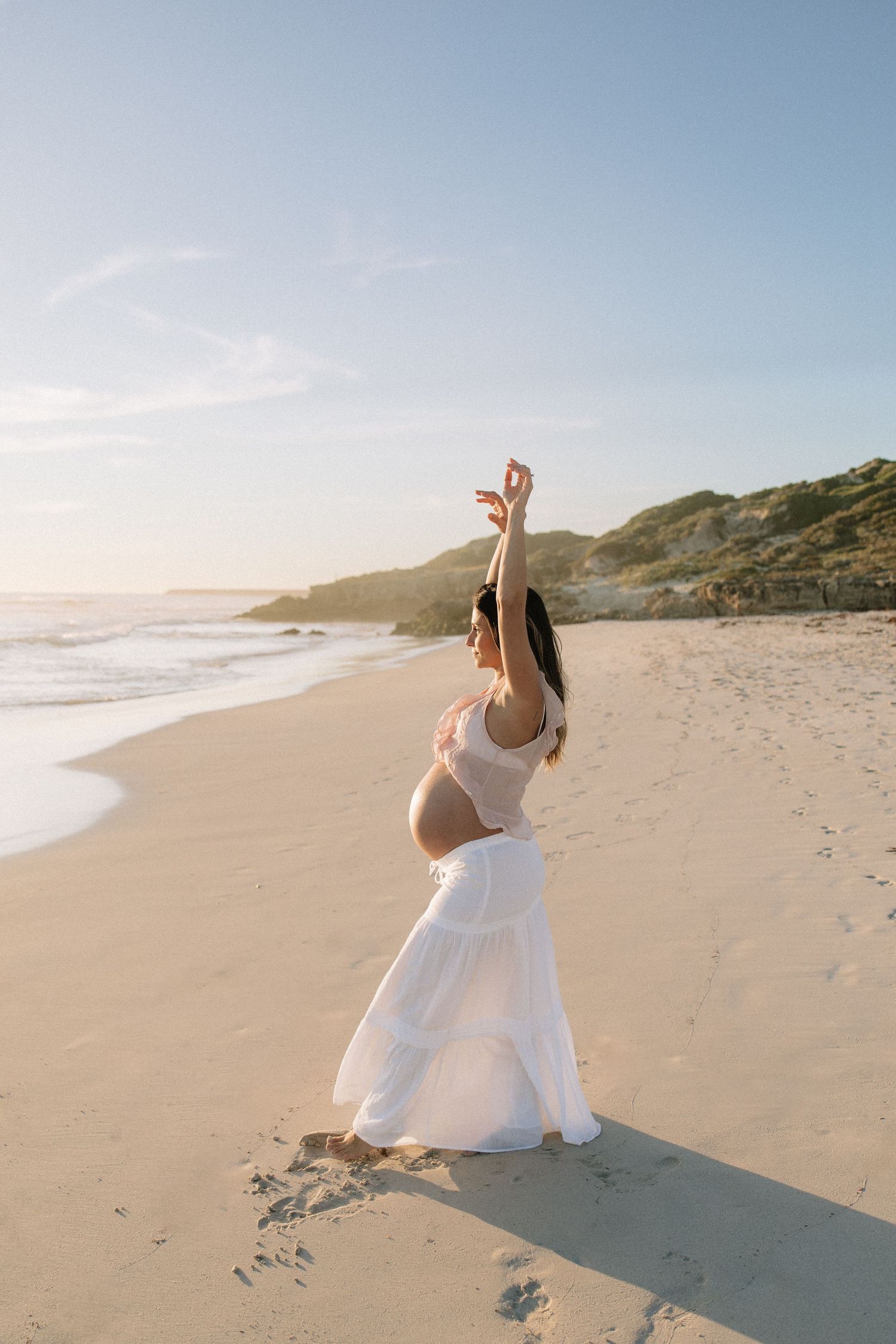 A pregnant woman in a white maxi skirt raises her arm gracefully on a sunlit beach at golden hour.