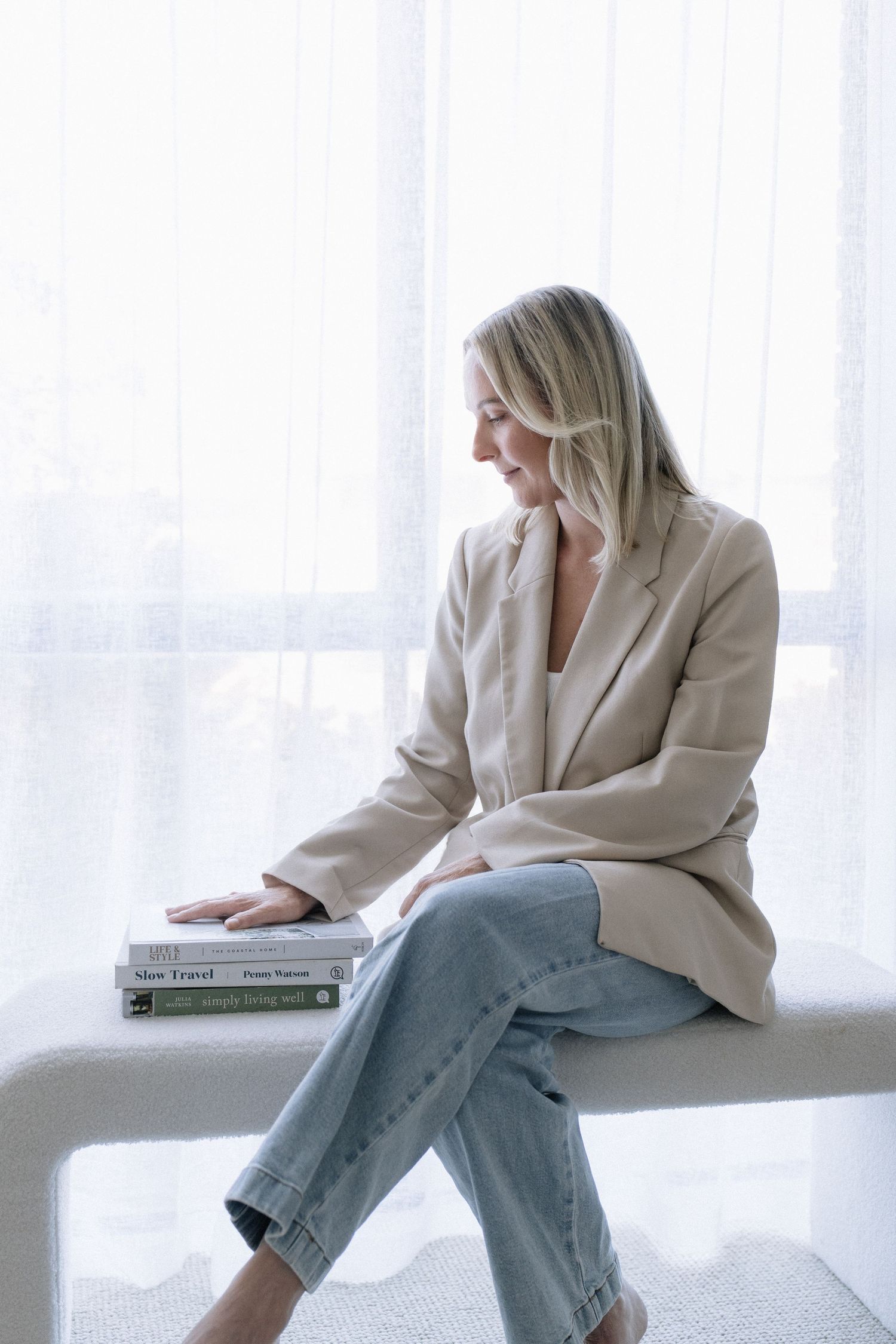 Stylish blonde woman in linen blazer and jeans sitting on white bench with books by sheer curtains.