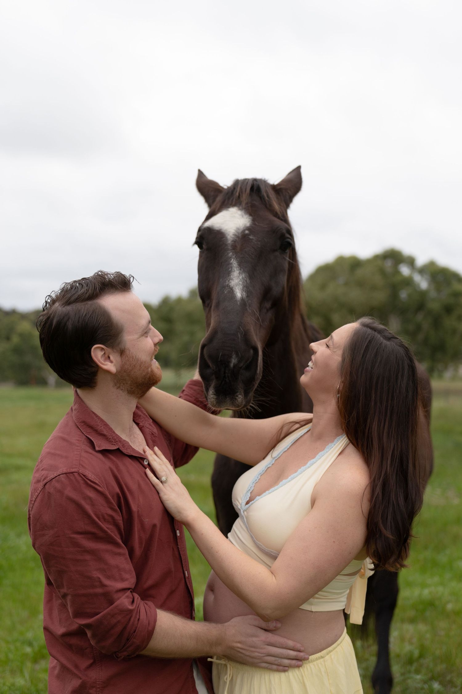 Smiling couple with pregnant woman petting a dark horse in a green field during maternity shoot.