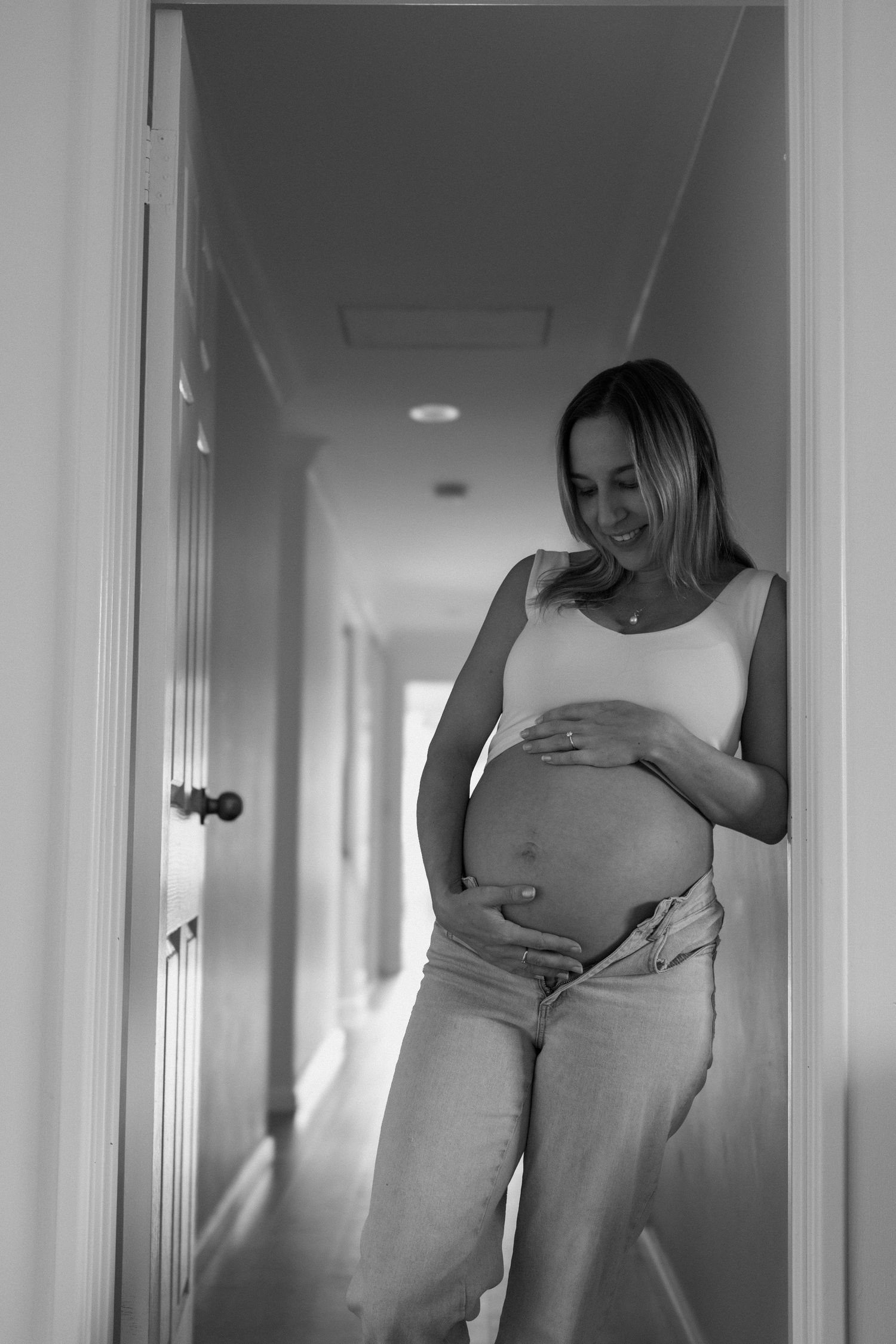 Smiling pregnant woman in white top and jeans holding baby bump in hallway, black and white photo.