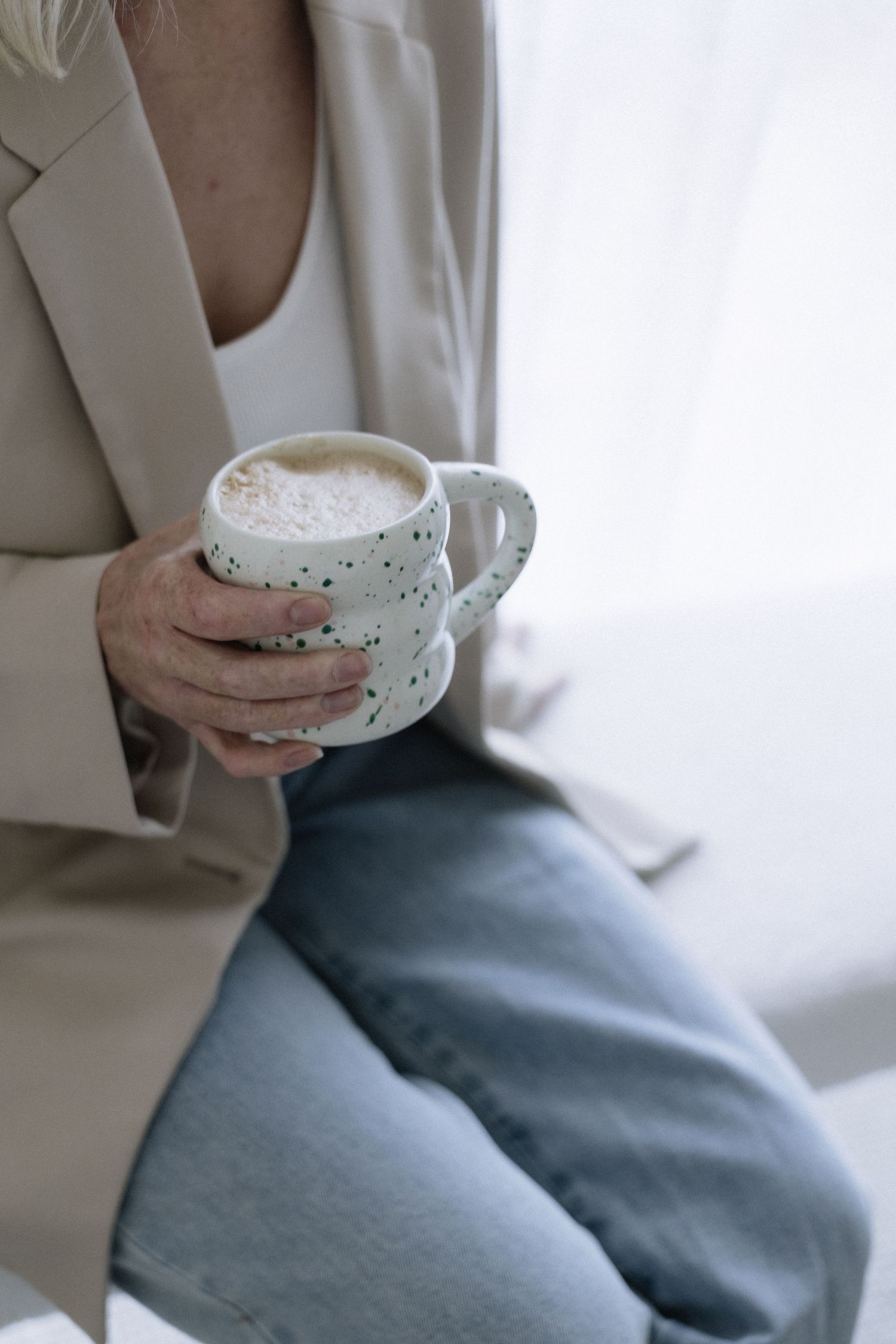 Close-up of woman in beige blazer and jeans holding speckled white ceramic coffee mug.