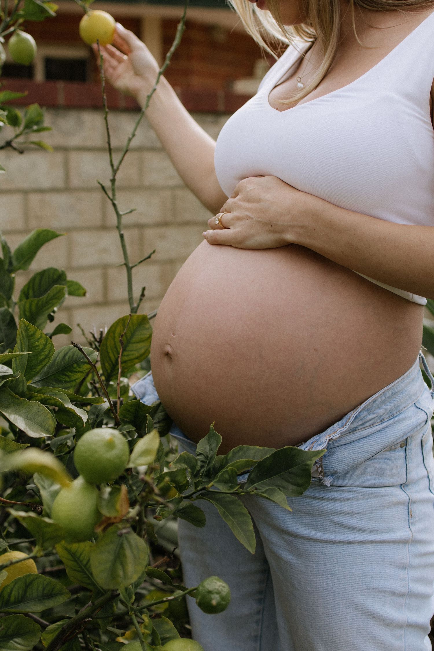 Pregnant woman in white top picking green apples from a tree while cradling her bare bump.