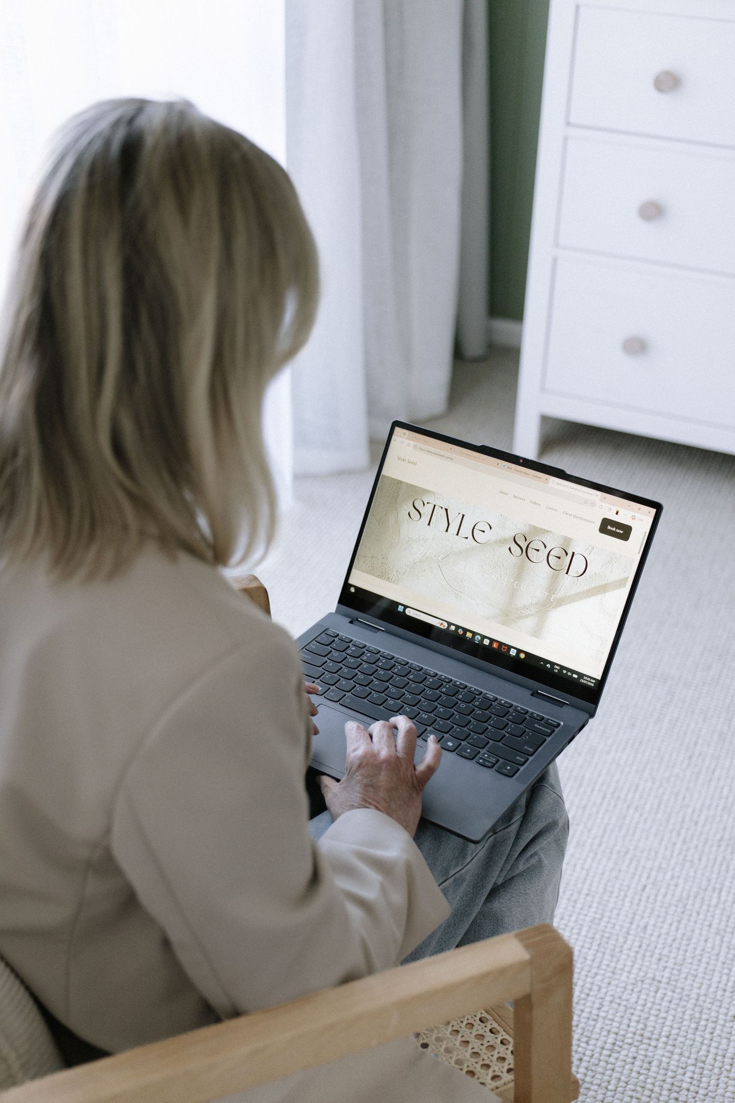 Blonde woman in beige blazer browsing Style Seed website on laptop while seated in rattan chair.