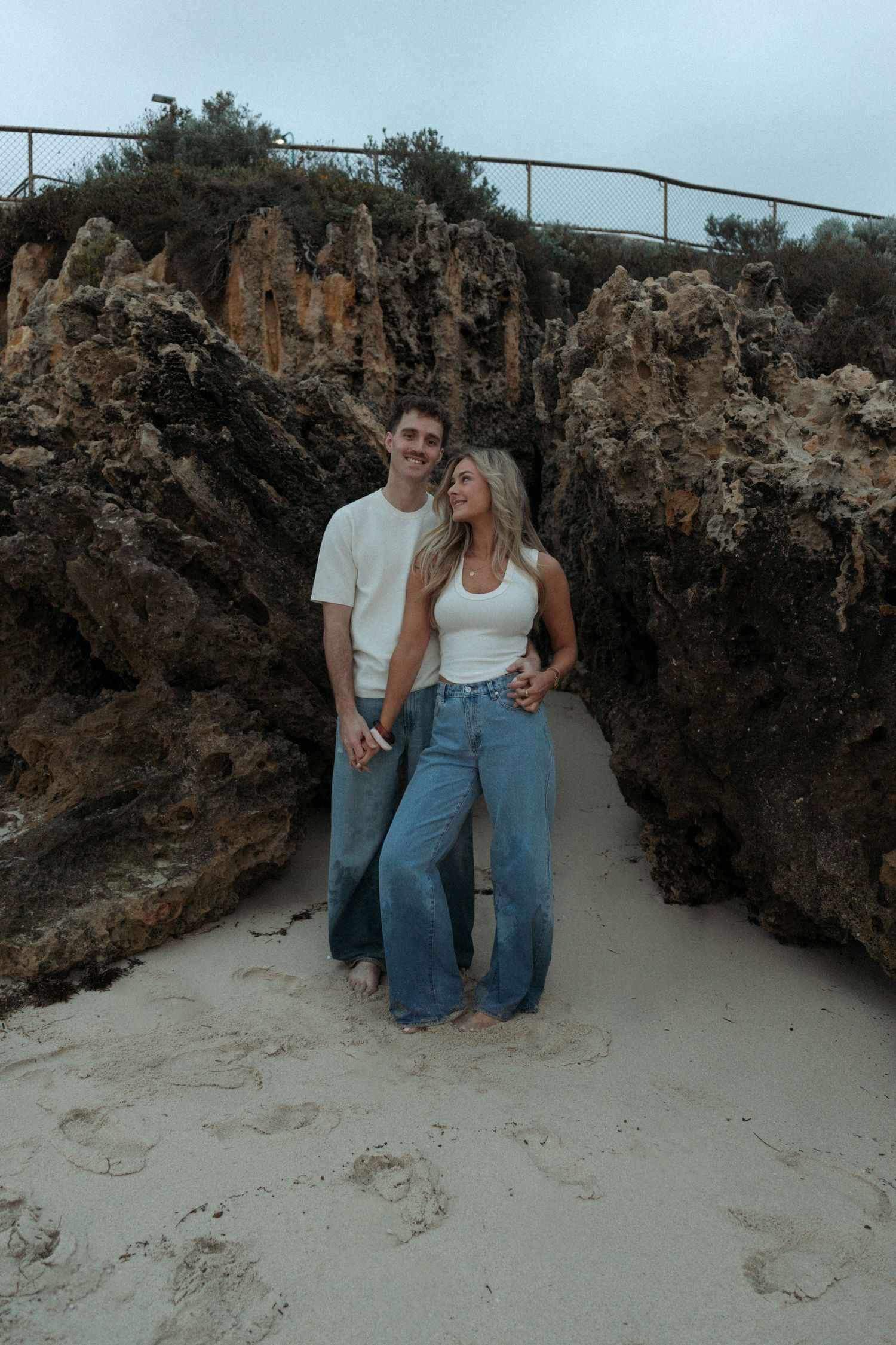 Smiling couple standing together between large rock formations on a sandy beach.